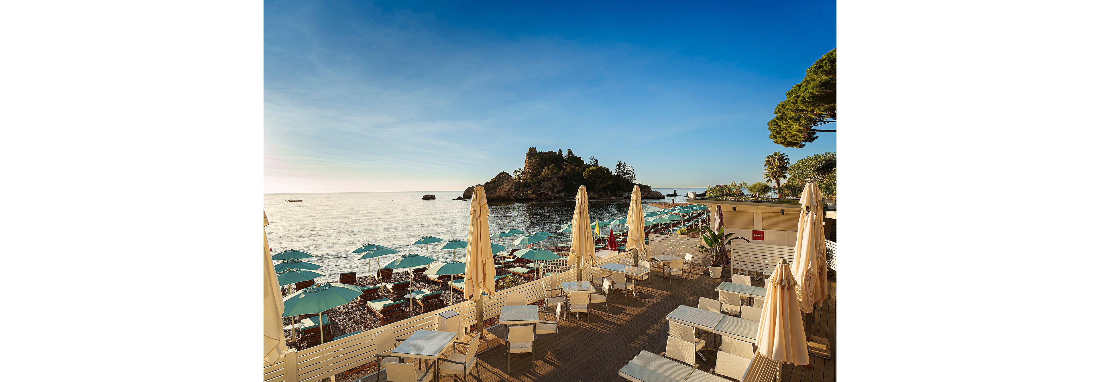 The main subject is a beachfront café featuring white tables and chairs, shaded by tall beige umbrellas. The setting is a sunny coastal area with turquoise beach umbrellas and lounge chairs arranged along the shore, facing a calm sea and a small rocky island in the distance. The visuals highlight a clear blue sky, tranquil water, and a relaxed vacation mood. No visible text or numeric values are present in the image.