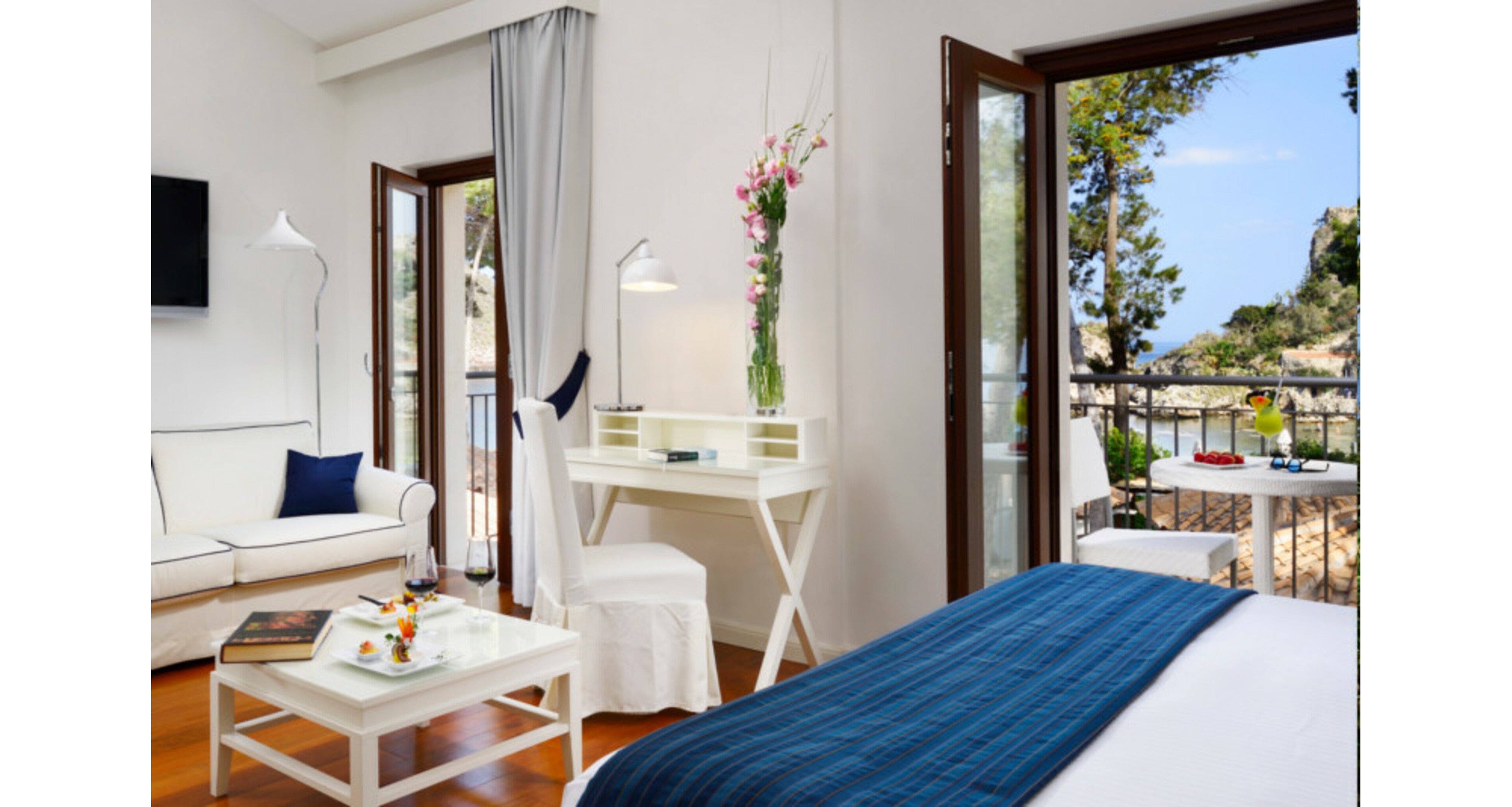 A contemporary hotel room features a white sofa, writing desk, and a bed with a blue throw. Large windows and open doors reveal a scenic balcony overlooking the sea and lush greenery. The space is decorated with fresh flowers, fruit, and books, creating a relaxing and inviting atmosphere.