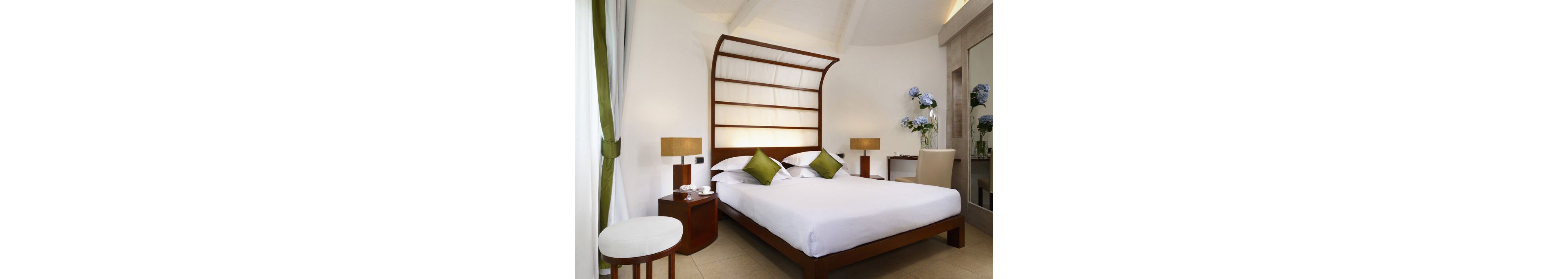 A contemporary hotel room features a wooden canopy bed with crisp white linens and green accent pillows. The space is decorated with matching bedside tables, lamps, and a small round table with a chair. Natural light filters through sheer curtains, and a vase of blue flowers sits on a desk in the background. The room has a clean, minimalist style with a neutral palette and green highlights.