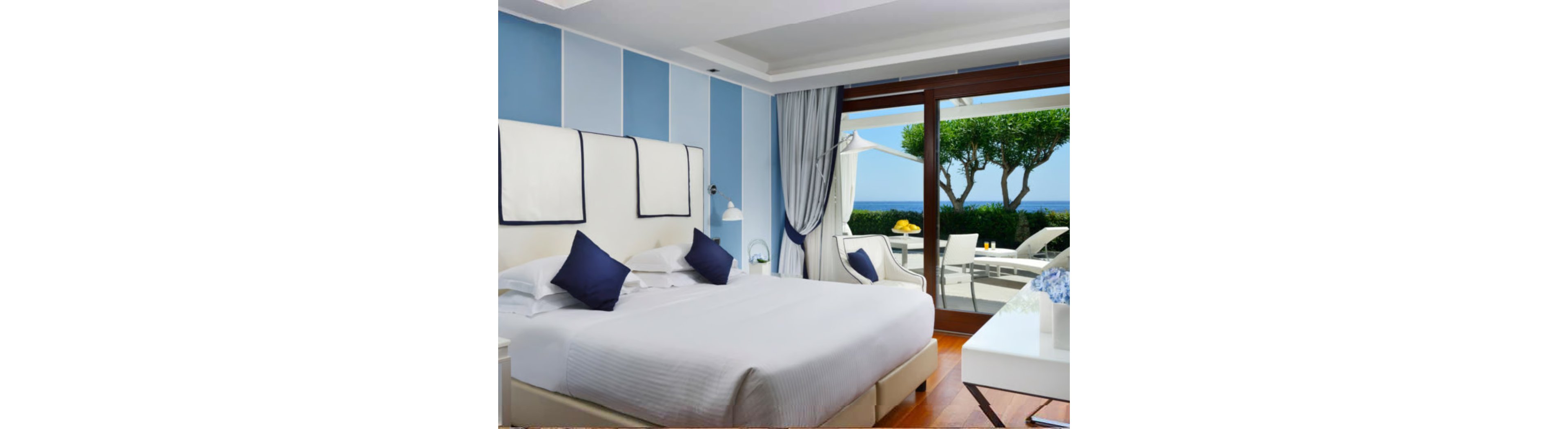 A stylish hotel room features a large bed with white linens and navy accent pillows, set against a light blue paneled wall. The room opens onto a private terrace with outdoor seating, overlooking the ocean and manicured greenery. Natural light fills the space, creating a serene and inviting atmosphere with a coastal color palette.