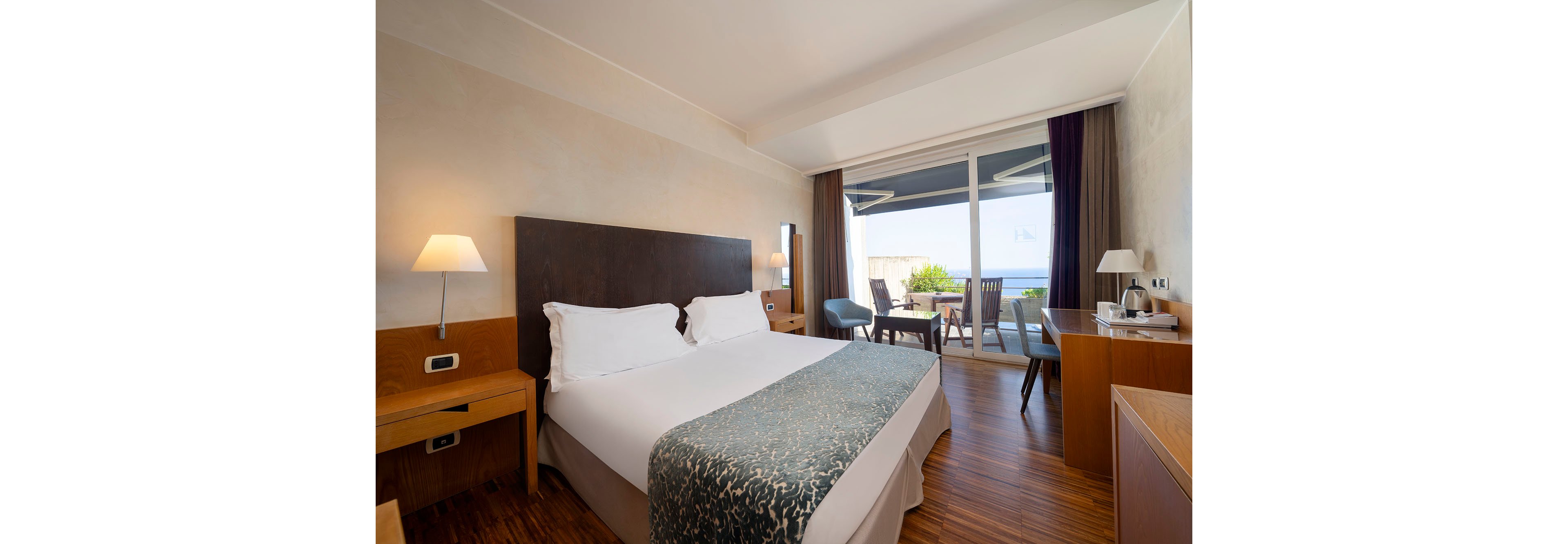 Bright contemporary hotel room features a double bed with white linens and a patterned bed runner. Wooden furniture, bedside lamps, and a desk create a warm and comfortable atmosphere. Large sliding glass doors open onto a balcony with chairs and a table overlooking the sea and greenery. The space is tidy and inviting, suitable for vacation or business travel.