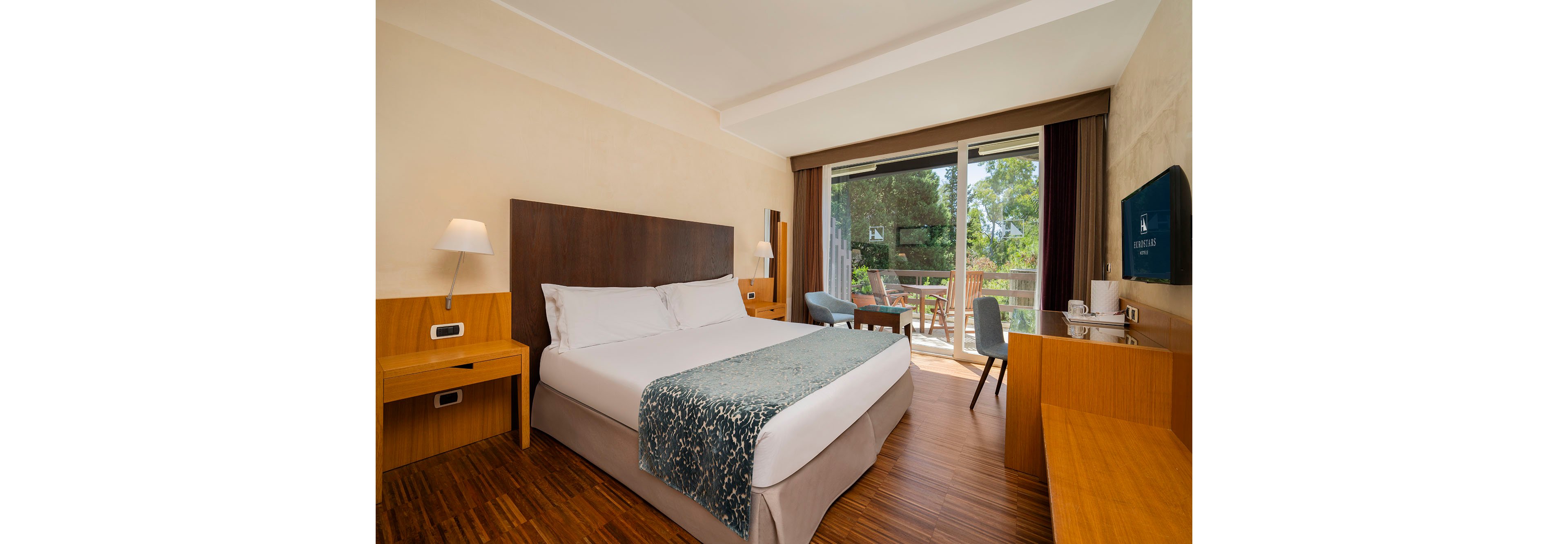 A modern hotel bedroom features a large double bed with white linens and a patterned teal throw. The room includes wooden bedside tables, a desk area, and a wall-mounted television. Floor-to-ceiling glass doors open onto a balcony with outdoor seating, overlooking lush green trees. Warm wood flooring and neutral walls create a clean, inviting atmosphere ideal for relaxation.