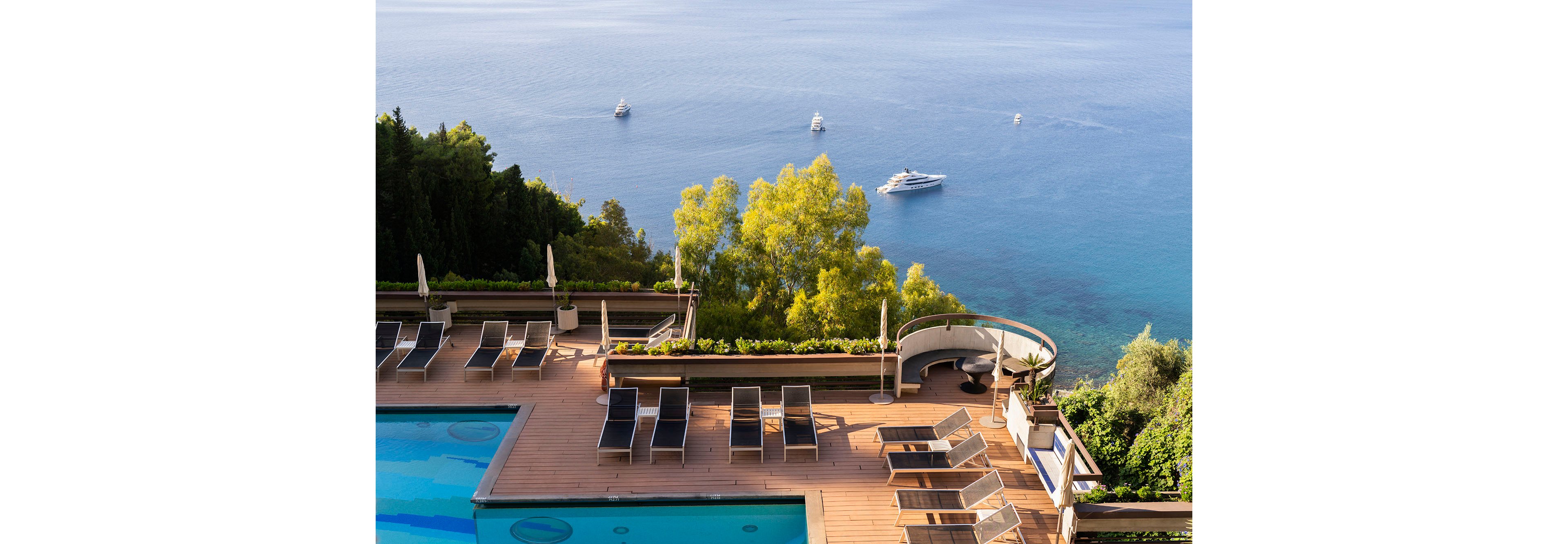 An elevated hotel pool terrace with sun loungers overlooks a calm blue sea dotted with several yachts. The foreground features a rectangular swimming pool and neatly arranged deck chairs on a wooden deck. Lush green trees and shrubs frame the terrace, creating a contrast with the deep blue water. The scene suggests a tranquil, upscale coastal resort atmosphere under clear daylight skies.