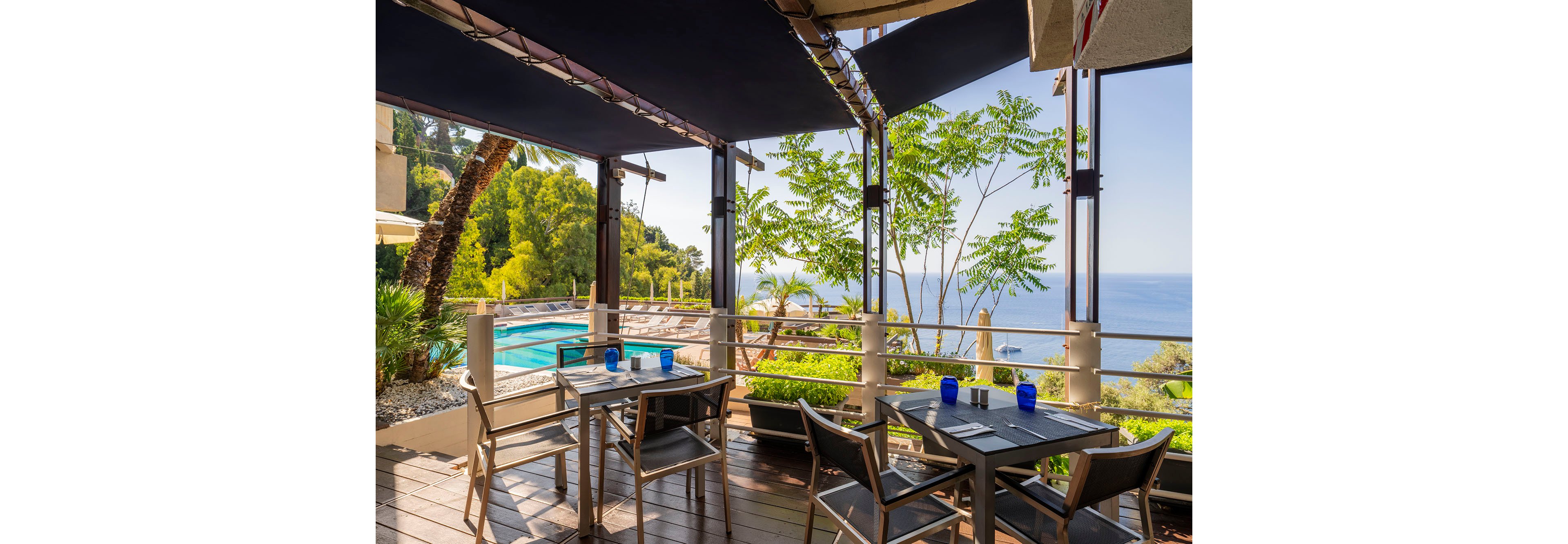 An outdoor terrace restaurant features several tables and chairs set with blue glass cups. The space is shaded by dark awnings and framed by railings and lush greenery. Below the terrace, a swimming pool sits among palm trees and landscaped plants. In the distance, calm blue sea and coastline stretch under a clear sunny sky, creating a relaxed vacation atmosphere.