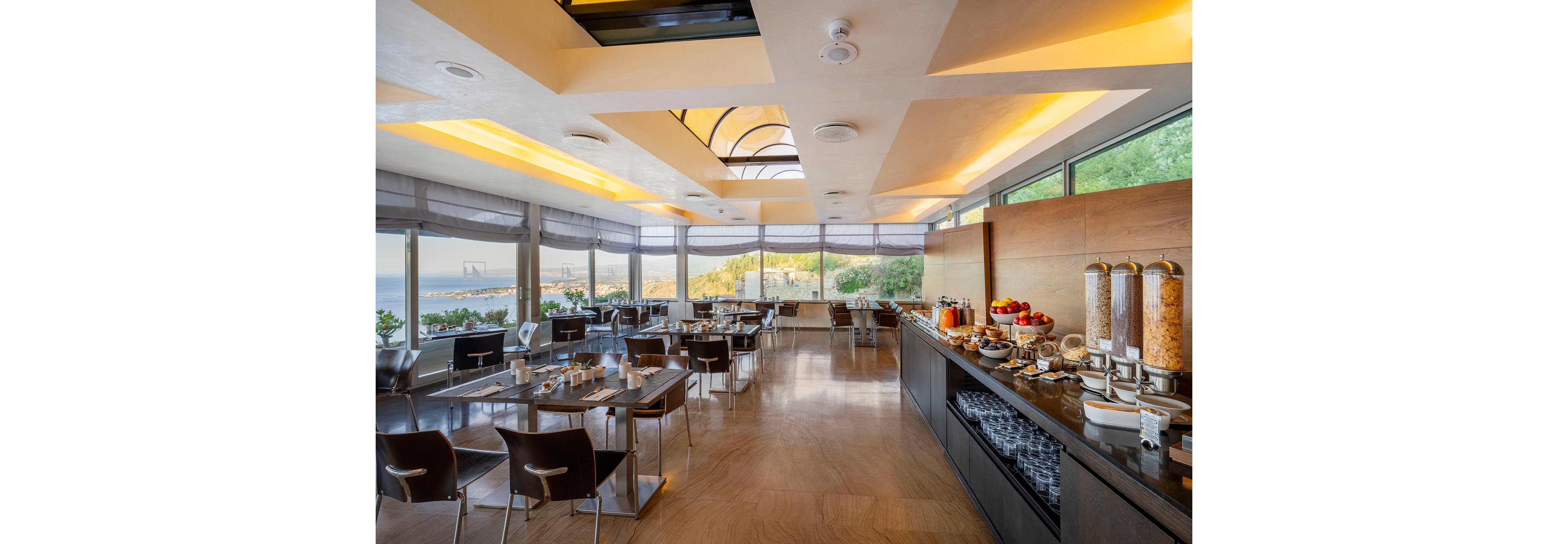 Spacious contemporary dining room featuring a self-service breakfast buffet. Large windows reveal a scenic coastal landscape and distant bridge, filling the space with natural light. The buffet counter displays cereals in dispensers, juices, fruits, and tableware neatly arranged. Sleek chairs and tables are set for guests, creating a clean, inviting atmosphere for morning dining.