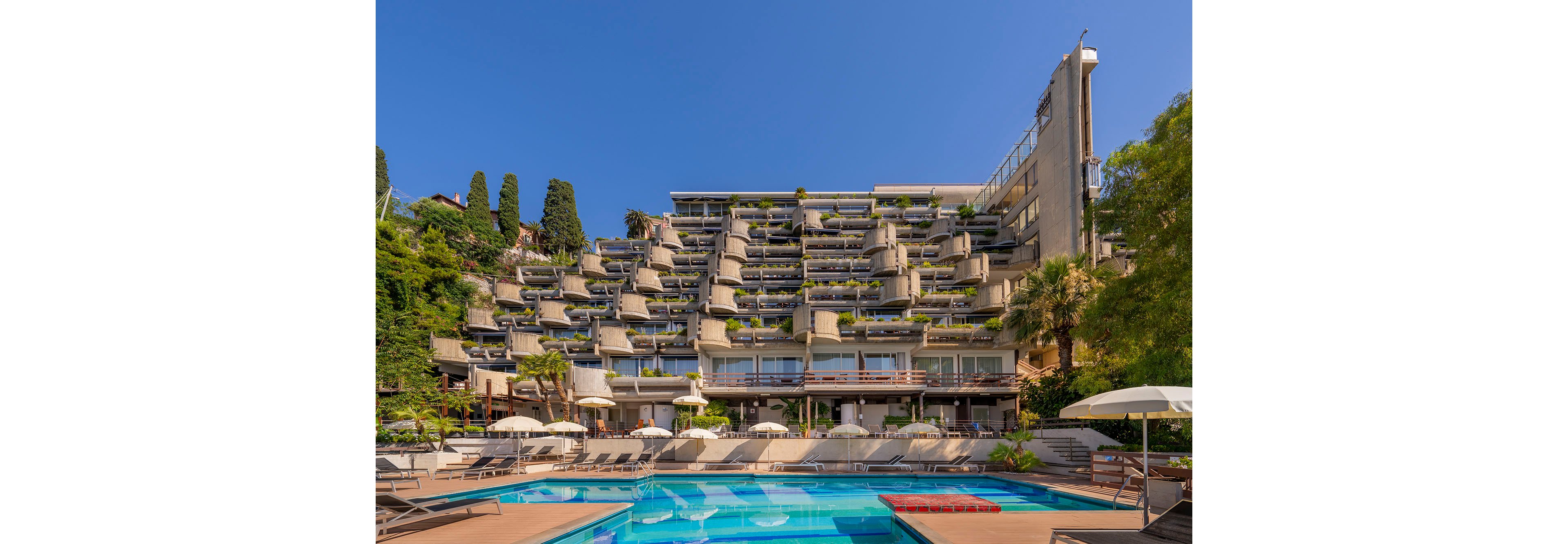 A large terraced hotel building rises above an outdoor swimming pool under a clear blue sky. The architecture features stacked balconies with greenery and plants, creating a stepped, modern facade. In the foreground, sun loungers and umbrellas line the pool deck, suggesting a relaxing vacation setting. Trees and lush vegetation frame the scene, adding a resort-like atmosphere.