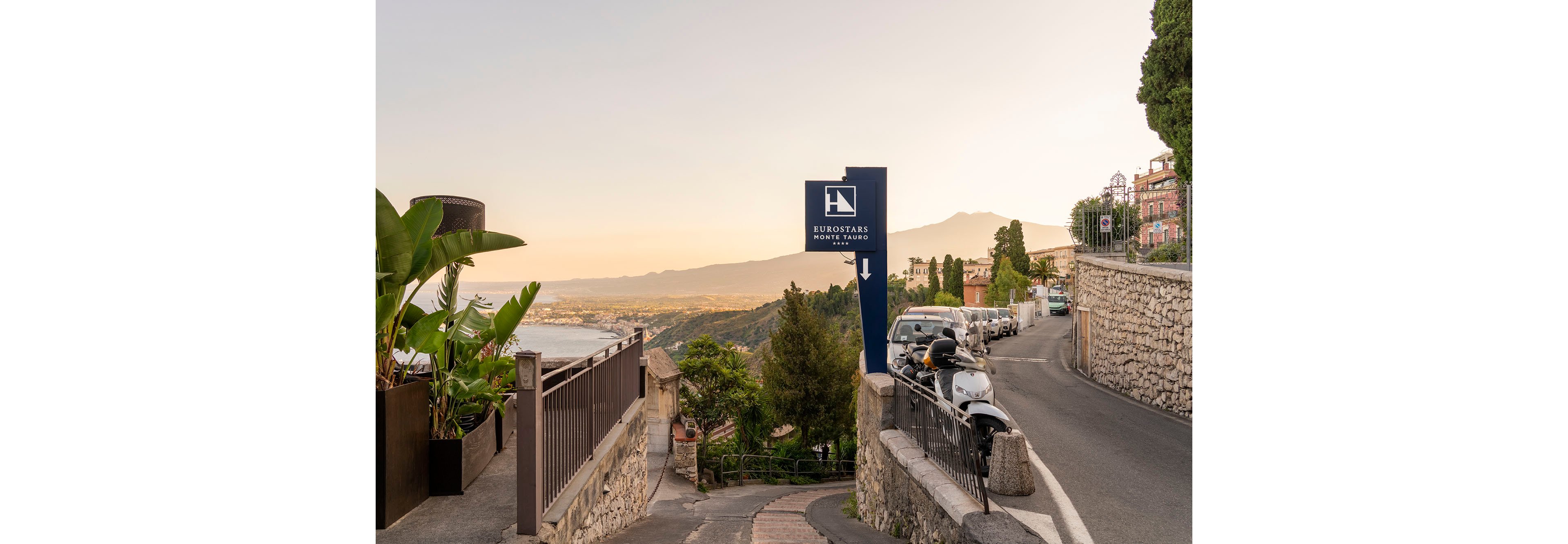 A narrow hillside street curves past a prominent Eurostars Hotels sign pointing downward. Parked cars and scooters line the road beside stone walls and railings. Lush plants and Mediterranean-style buildings frame the view, leading the eye toward a distant coastline and mountains at sunset. Soft golden light creates a calm, scenic atmosphere in this urban landscape.
