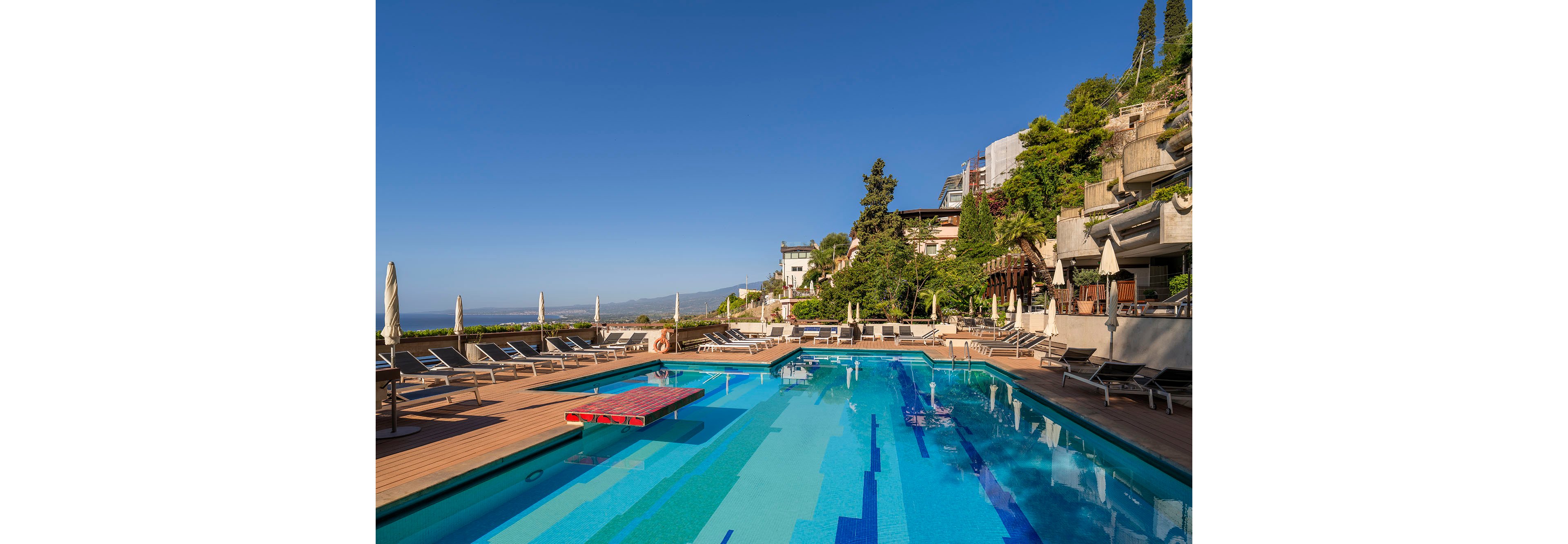 An outdoor swimming pool sits on a wooden deck at a hillside hotel overlooking the sea. Sun loungers and closed umbrellas line the poolside, suggesting a tranquil resort setting. Terraced buildings and lush greenery rise along the slope, adding architectural interest and a Mediterranean feel. The clear blue sky and calm water create a relaxing vacation atmosphere.