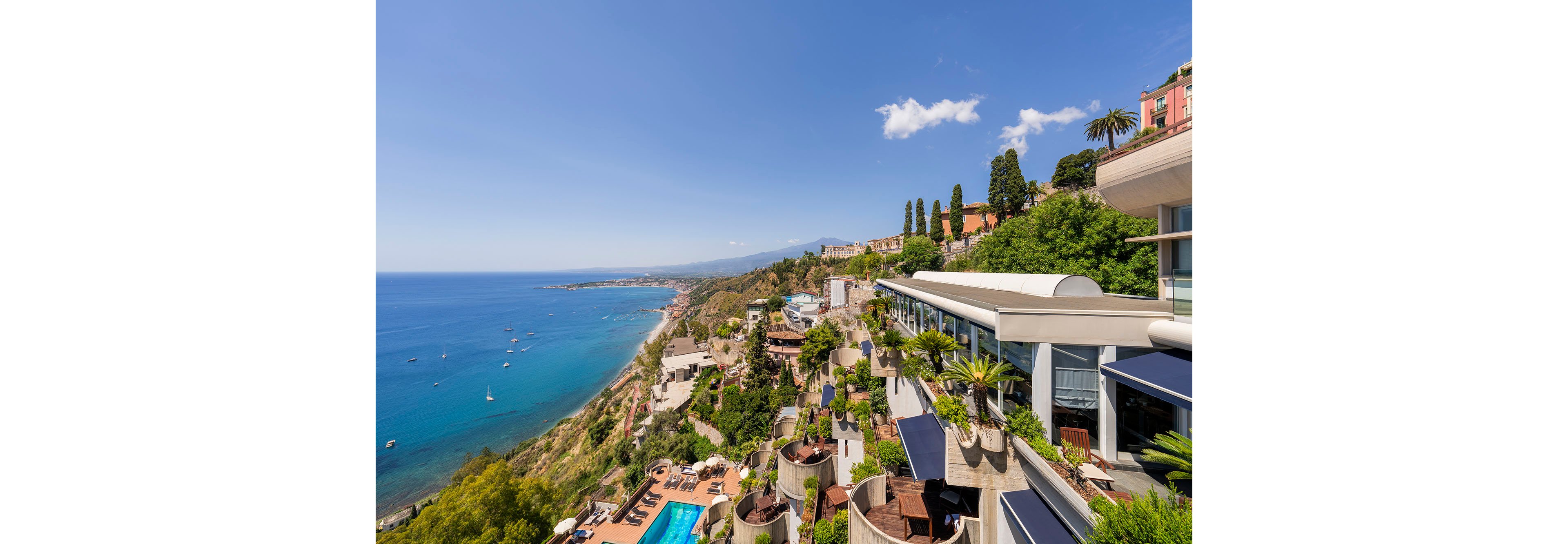 A modern hillside resort stretches along a bright blue coastline under a clear sunny sky. Terraced buildings with glass facades and lush greenery descend toward the water, featuring an outdoor swimming pool and sun loungers. The expansive sea view shows boats scattered across the calm bay and a distant shoreline curving into the horizon. Tall cypress trees and colorful villas add a picturesque touch to the scenic coastal landscape.