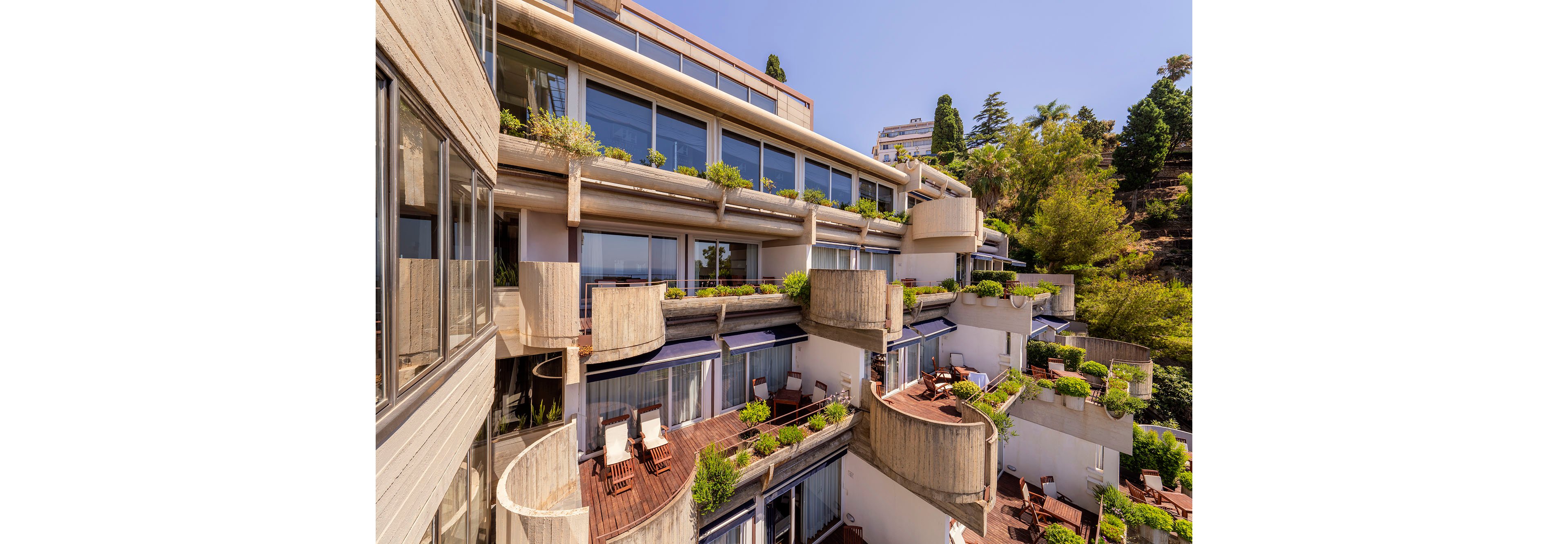 A multi-level modern building features numerous terraced balconies overlooking a hillside. Each balcony has wooden decking, outdoor lounge chairs, and potted plants that create a lush, inviting atmosphere. Large glass windows line the façade, emphasizing contemporary architecture and open views. Trees and vegetation surround the structure, enhancing the resort-like, sunny setting.