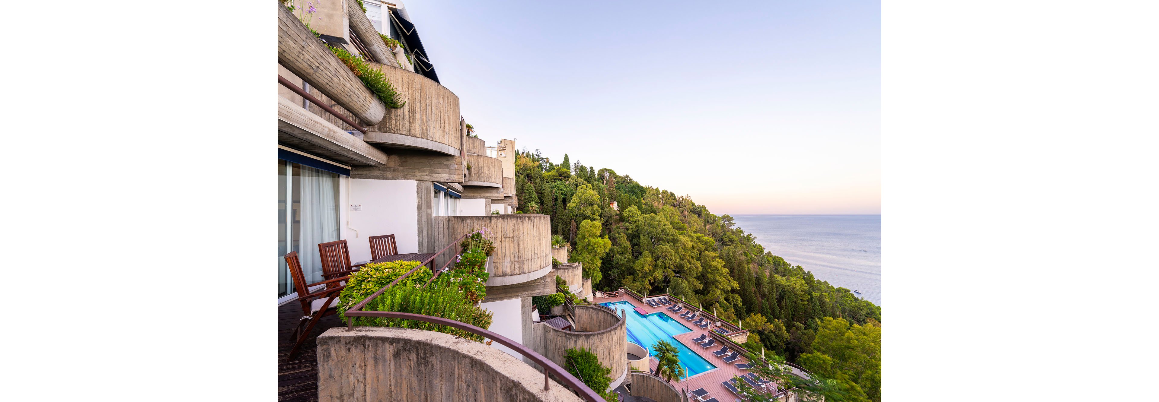 A modern terraced hotel overlooks a large outdoor swimming pool and lush green hillside by the sea. Wooden chairs and balconies create an inviting space for guests to relax and enjoy the coastal view. The scene is captured at sunset or early evening, with soft pastel light over the calm water. The composition emphasizes the layered architecture, vibrant trees, and tranquil resort atmosphere.