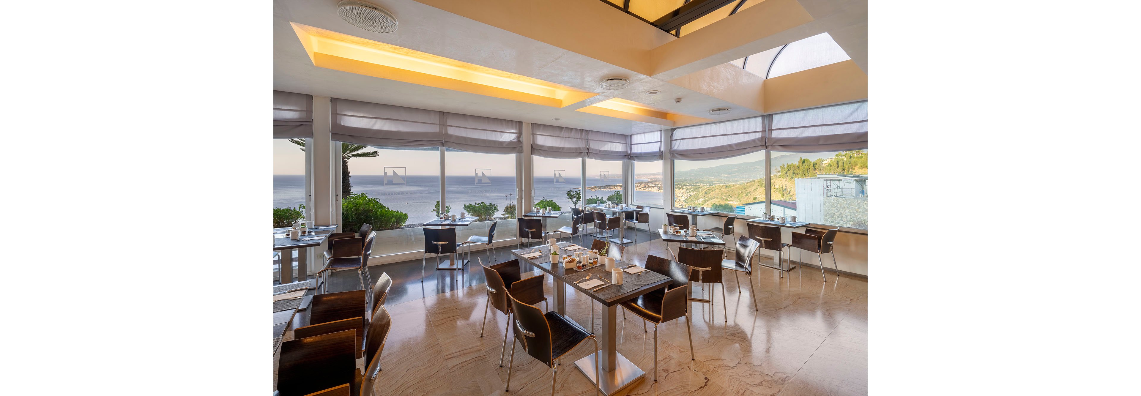 Bright contemporary restaurant interior features neatly arranged tables and chairs facing large windows. The space overlooks a scenic coastline and expansive ocean, with hills and greenery visible in the distance. Morning or daytime light fills the room, highlighting clean surfaces and minimalist decor. Table settings with cups, plates, and condiments suggest a relaxed dining experience with a view.