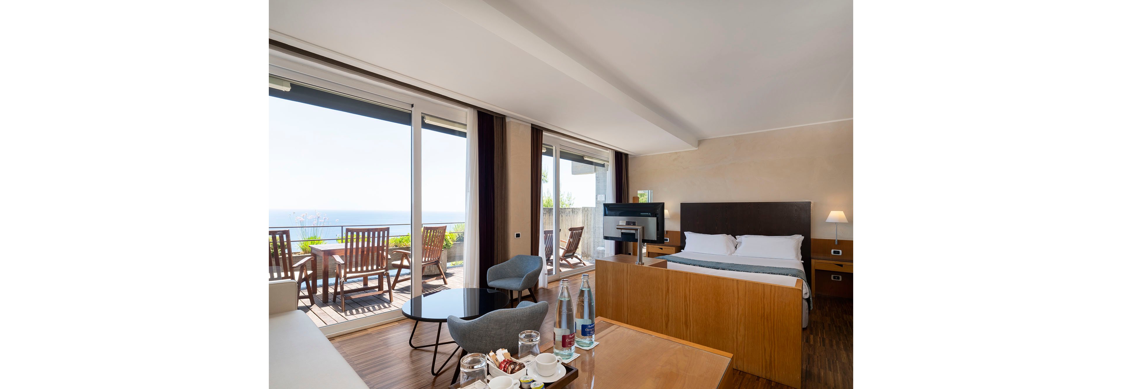 A contemporary hotel room features a large bed, seating area, and desk facing a flat-screen television. Floor-to-ceiling glass doors open onto a wooden terrace with outdoor chairs and a table overlooking the ocean. Inside, a round coffee table holds glassware and bottled water, while a tray with cups and snacks sits on the desk. Warm neutral tones, wood flooring, and soft lighting create a relaxed, upscale atmosphere.