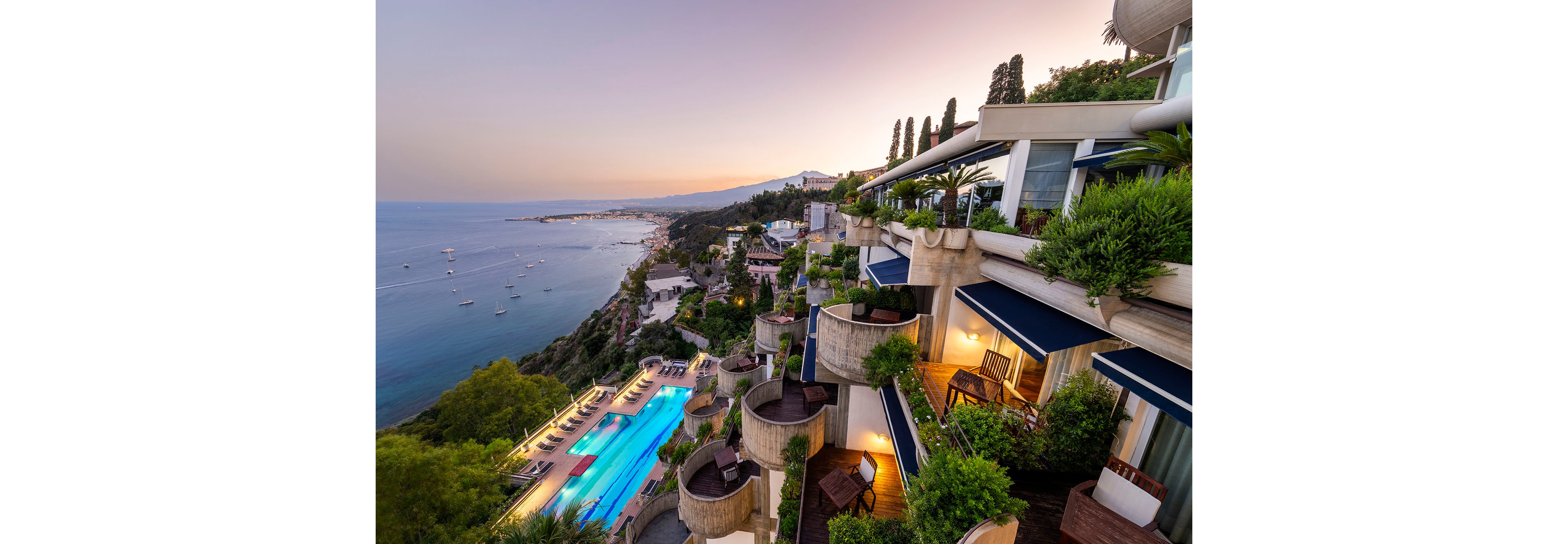 A luxury terraced resort overlooks a calm coastal bay at sunset. Multiple balconies with outdoor seating and lush greenery cascade down the hillside toward an illuminated infinity-style swimming pool. Sun loungers line the pool deck, while sailboats and yachts dot the tranquil sea in the distance. The warm evening sky and coastal landscape create a serene, upscale vacation atmosphere.