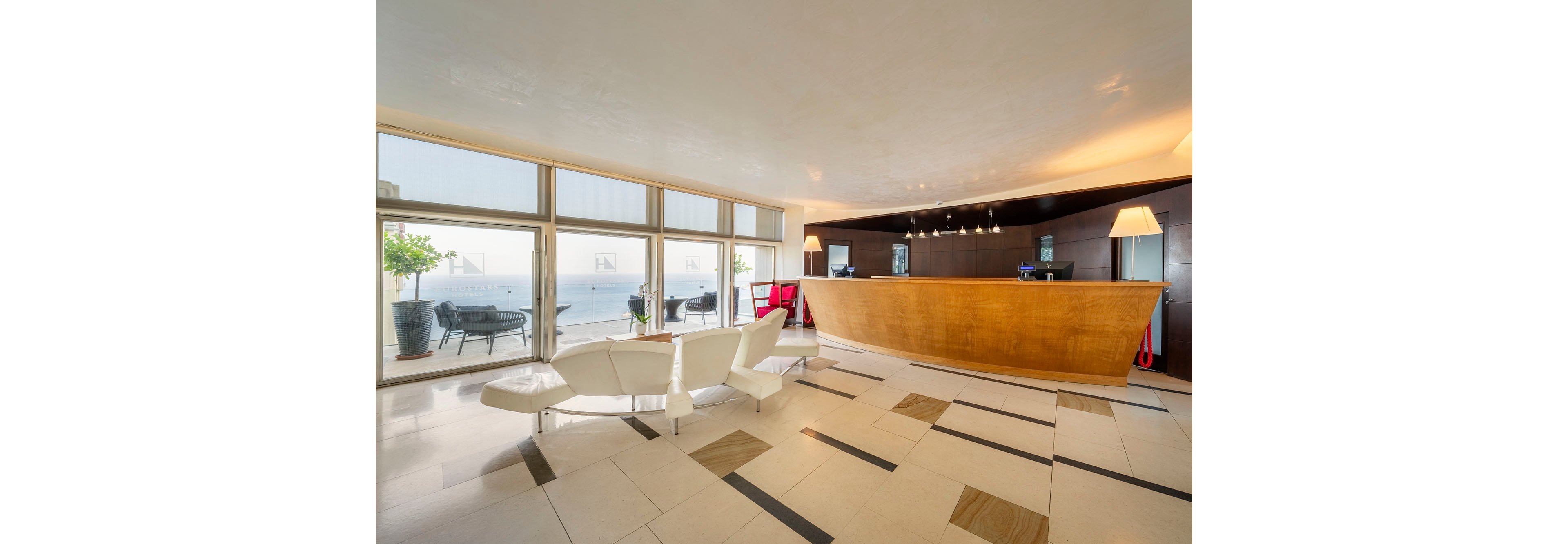 A contemporary hotel reception area features a large wooden front desk and sleek white seating. Floor-to-ceiling windows reveal a bright oceanfront view with outdoor seating and potted plants. The interior has polished tile flooring with geometric patterns and warm ambient lighting from table lamps and ceiling fixtures. The space appears calm and welcoming, designed for guests checking in or relaxing near the lobby.