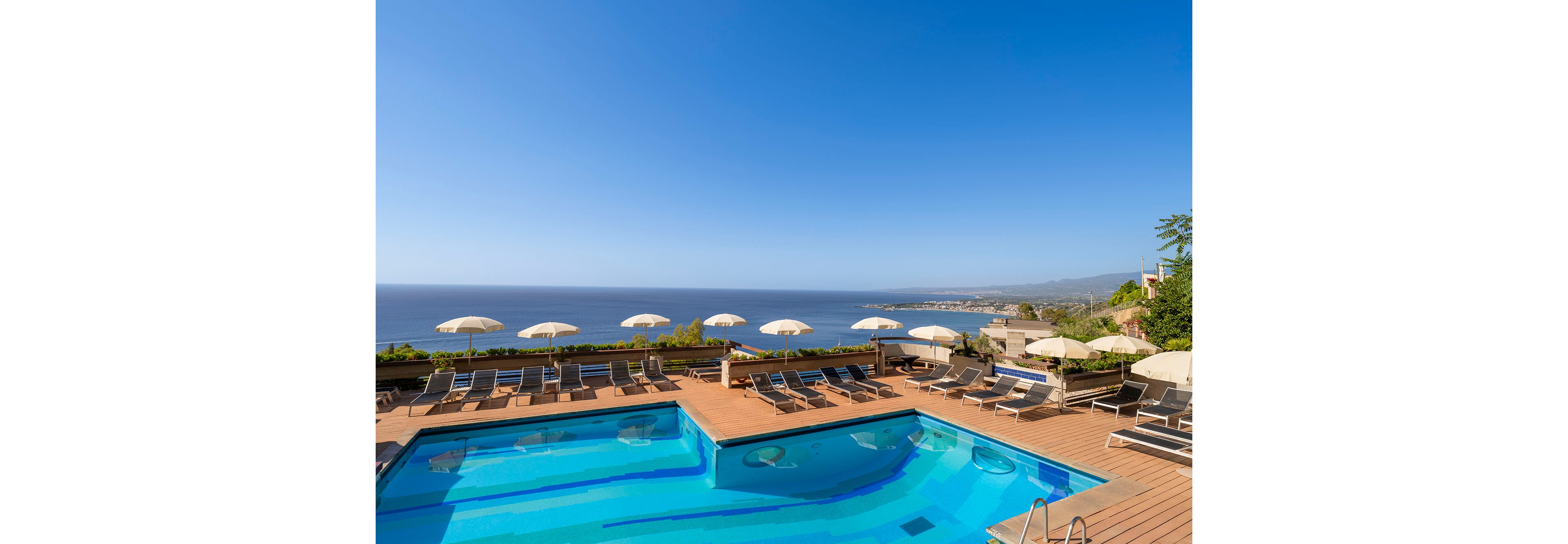 An outdoor hotel pool sits on a wooden terrace with multiple sun loungers arranged around it. Cream-colored umbrellas line the edge of the deck, facing a wide expanse of calm blue ocean. The sky is clear and bright, creating a warm, inviting atmosphere. In the distance, coastal buildings and hills are visible along the shoreline.