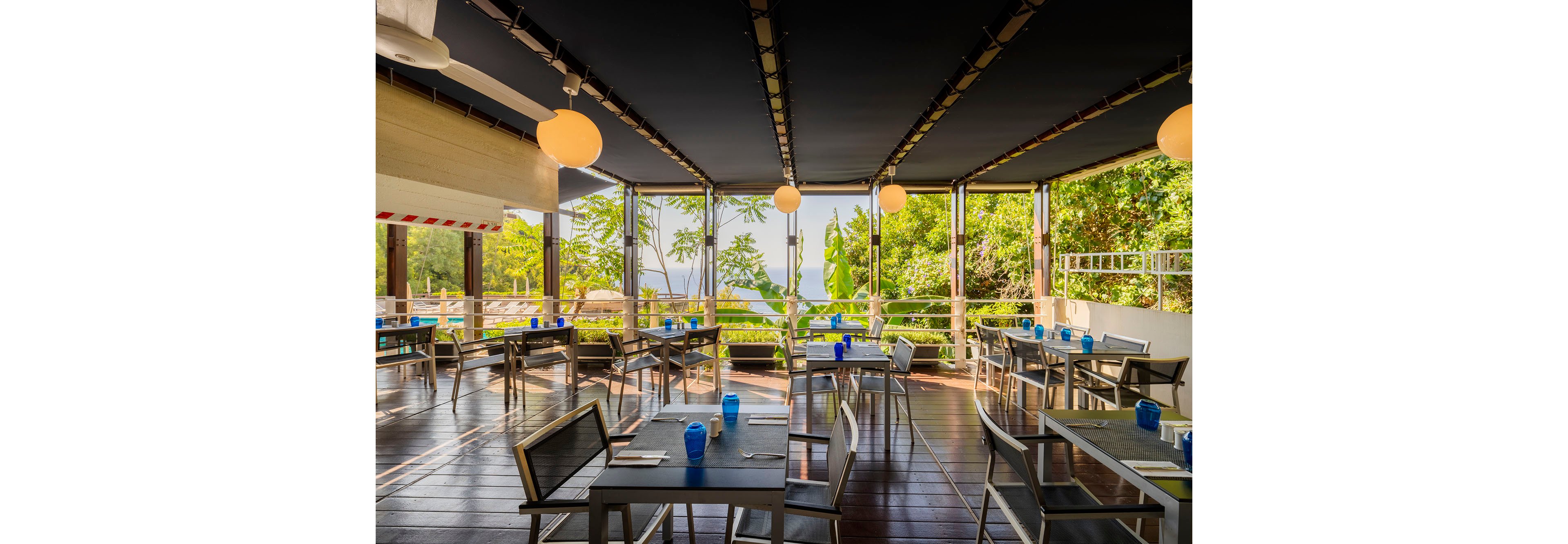 An open-air restaurant terrace features neatly arranged tables and chairs with blue glasses and simple place settings. The space is covered by a dark canopy and framed by metal railings and posts, creating a modern, airy dining area. Large round hanging lights and ceiling fans add comfort and ambiance. Beyond the terrace, dense green foliage and a distant view of water suggest a tropical or coastal location.