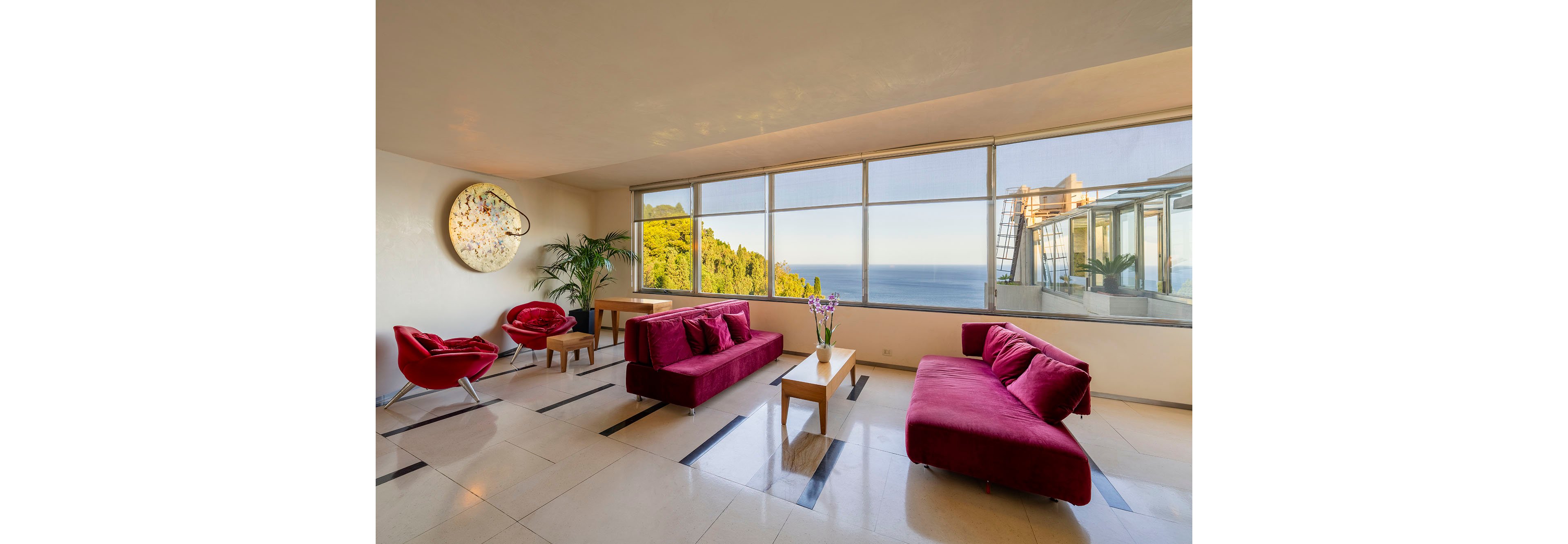 A contemporary living room features bold red sofas and armchairs arranged around light wood coffee tables. Large floor-to-ceiling windows reveal a scenic ocean view and lush green hillside outside. The interior is bright and minimal, with polished tile flooring and a decorative circular wall art piece. Natural light fills the space, creating an airy and inviting atmosphere.