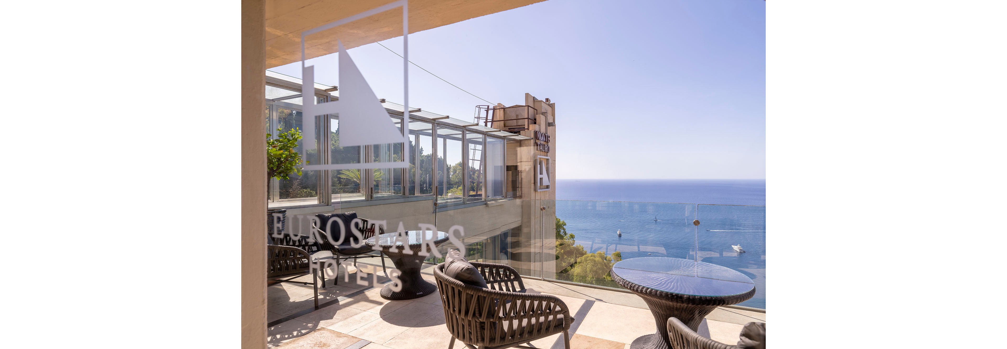 A modern hotel terrace overlooks a calm blue sea under a clear sky. Wicker chairs and a round table are arranged near a glass railing, creating a relaxed outdoor seating area. The hotel structure and elevator tower rise beside the terrace, with the words "EUROSTARS HOTELS" and other signage visible on the glass and building. Boats can be seen in the water below, enhancing the coastal vacation atmosphere.