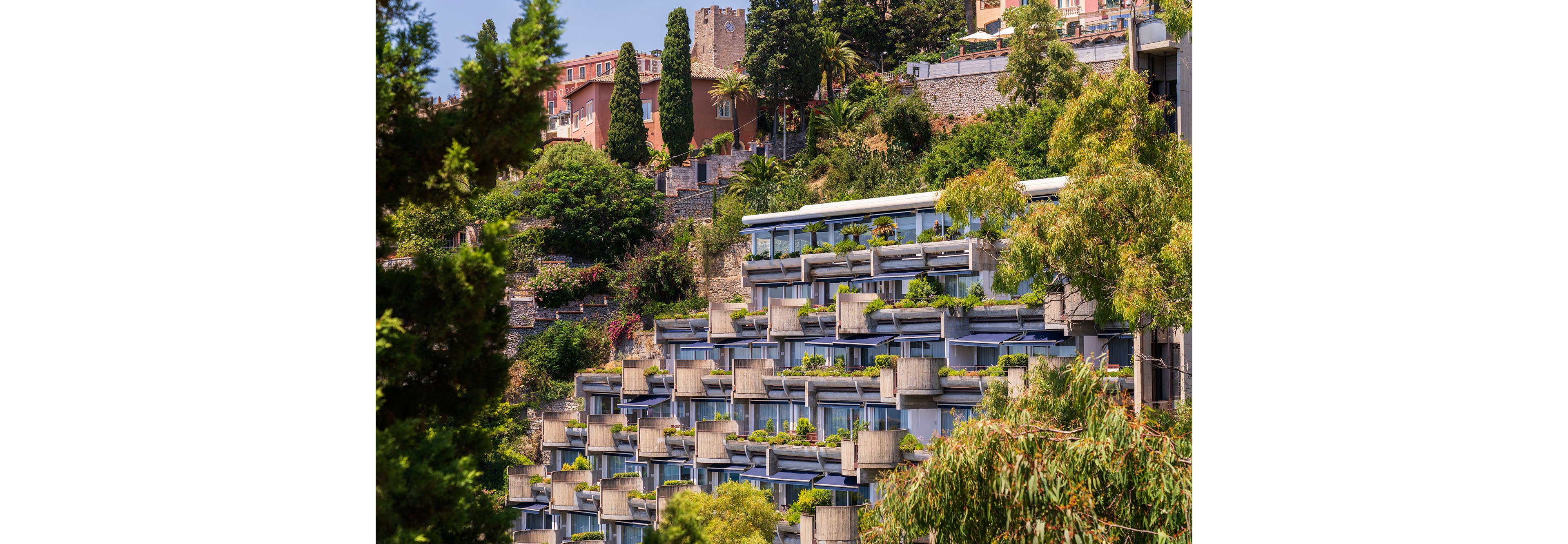 The image shows a series of modern terraced apartment buildings built into a steep hillside. Each level features concrete balconies filled with lush green plants and shrubs. Behind the contemporary structure, older houses and stone walls rise among tall cypress trees and other vegetation. The scene suggests a sunny Mediterranean or coastal town with abundant greenery and layered architecture.