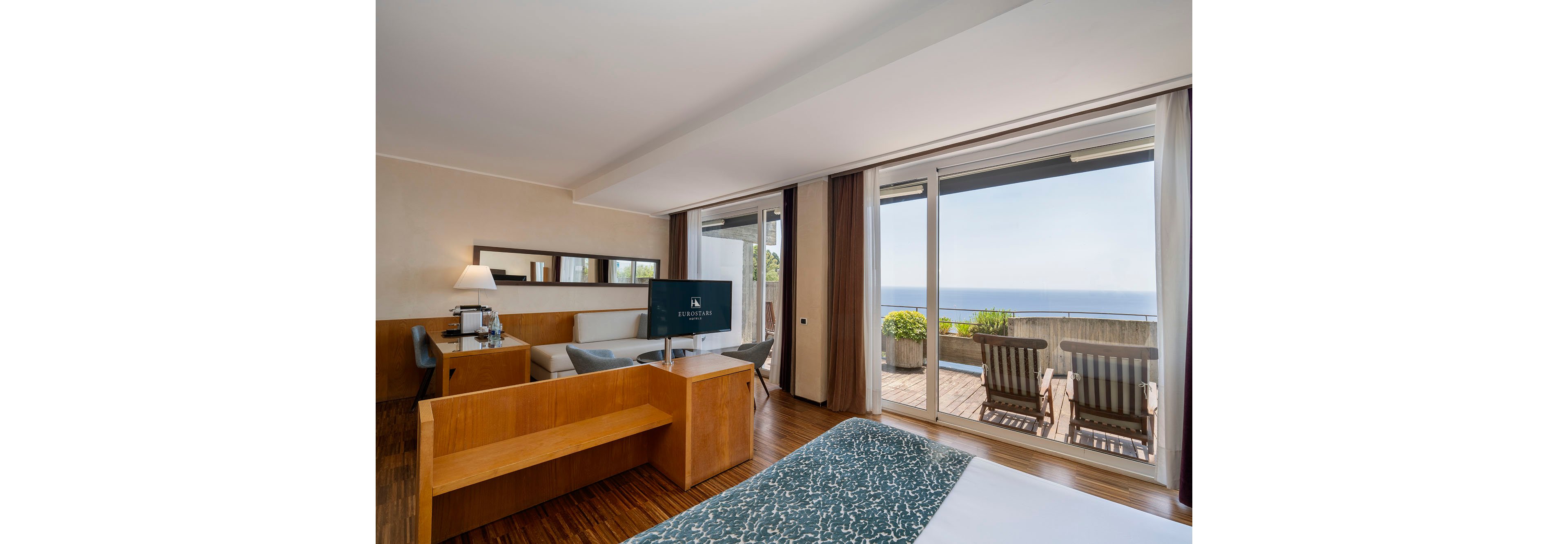 A contemporary hotel room features a neatly made bed, a wooden desk, and a seating area with a sofa and armchair. Large floor-to-ceiling glass doors open onto a private terrace with wooden chairs and a table overlooking the ocean. Neutral walls and warm wood flooring create a calm, inviting atmosphere, complemented by soft natural light. The scene suggests a relaxing vacation or business stay in a coastal destination.
