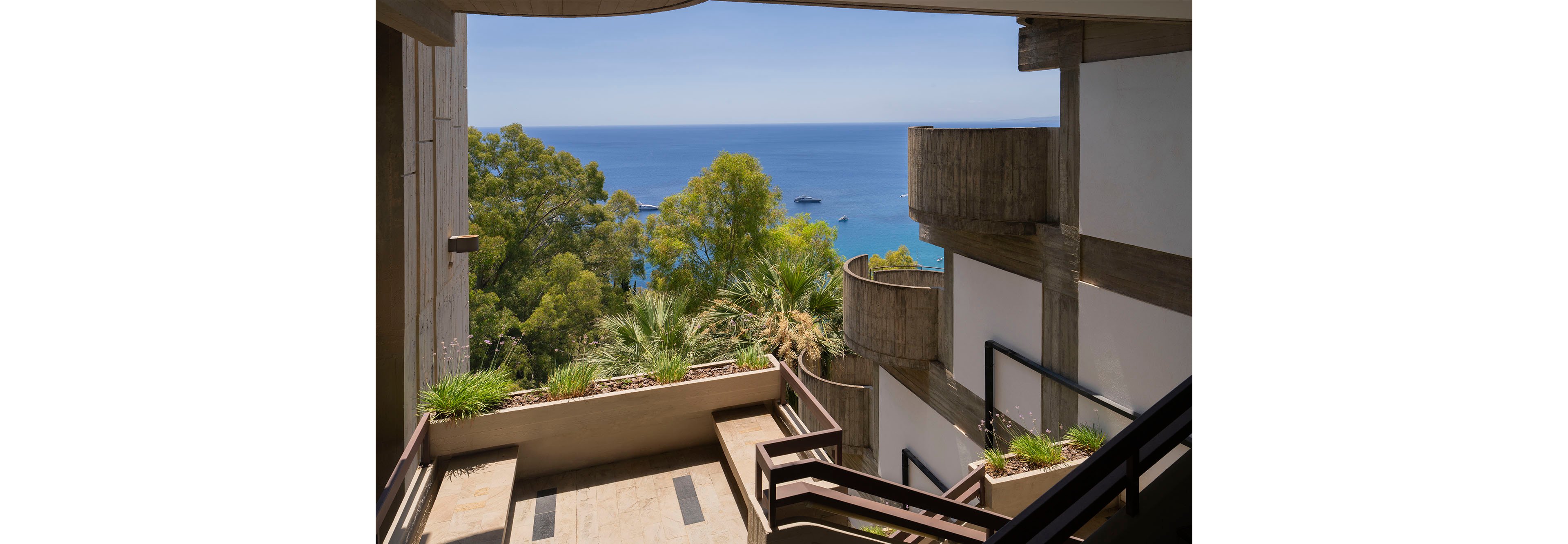A concrete terrace staircase descends through a modern building toward a scenic coastal view. Lush green trees and palm plants frame the architecture, softening the angular lines of the structure. Beyond the terrace, the calm blue sea stretches to the horizon under a clear sky, with small boats visible in the distance. The image combines brutalist architectural elements with natural seaside scenery for a tranquil, contemporary atmosphere.