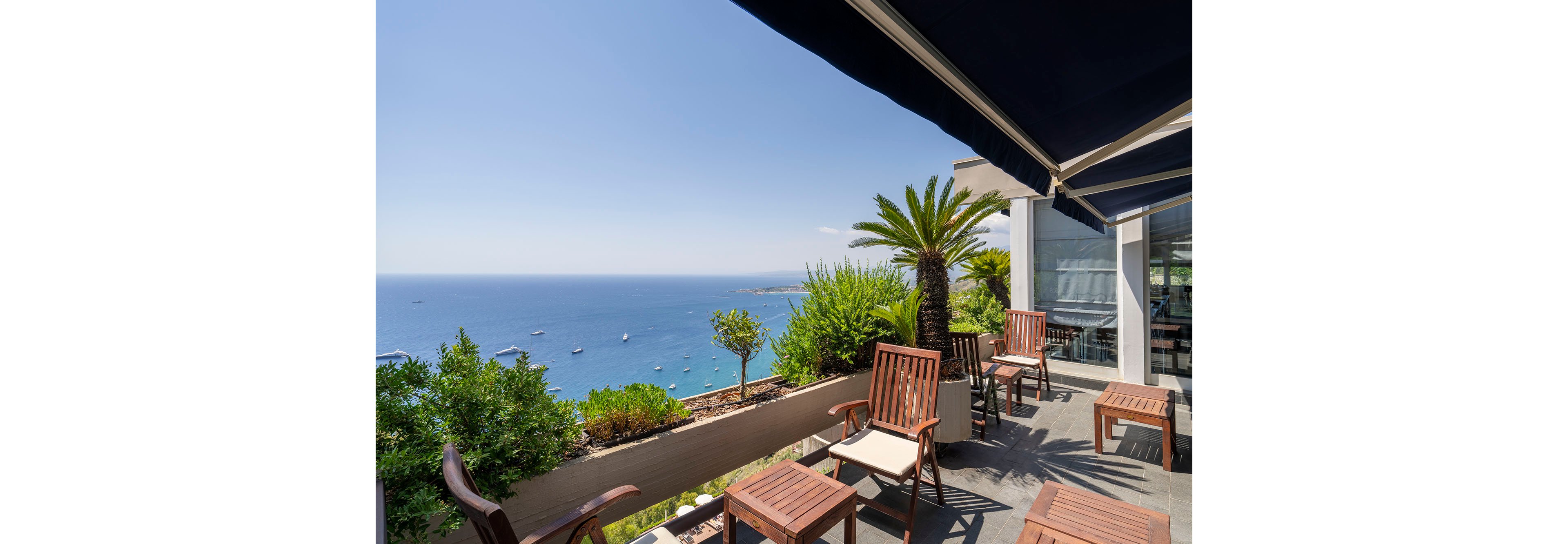 An outdoor terrace overlooks a calm blue sea dotted with boats under a clear sky. Wooden chairs and tables are arranged along the edge, shaded by a dark awning. Lush green plants and a palm tree line the balcony, adding a tropical touch to the setting. The scene suggests a relaxing coastal resort or café environment with bright daylight and tranquil atmosphere.