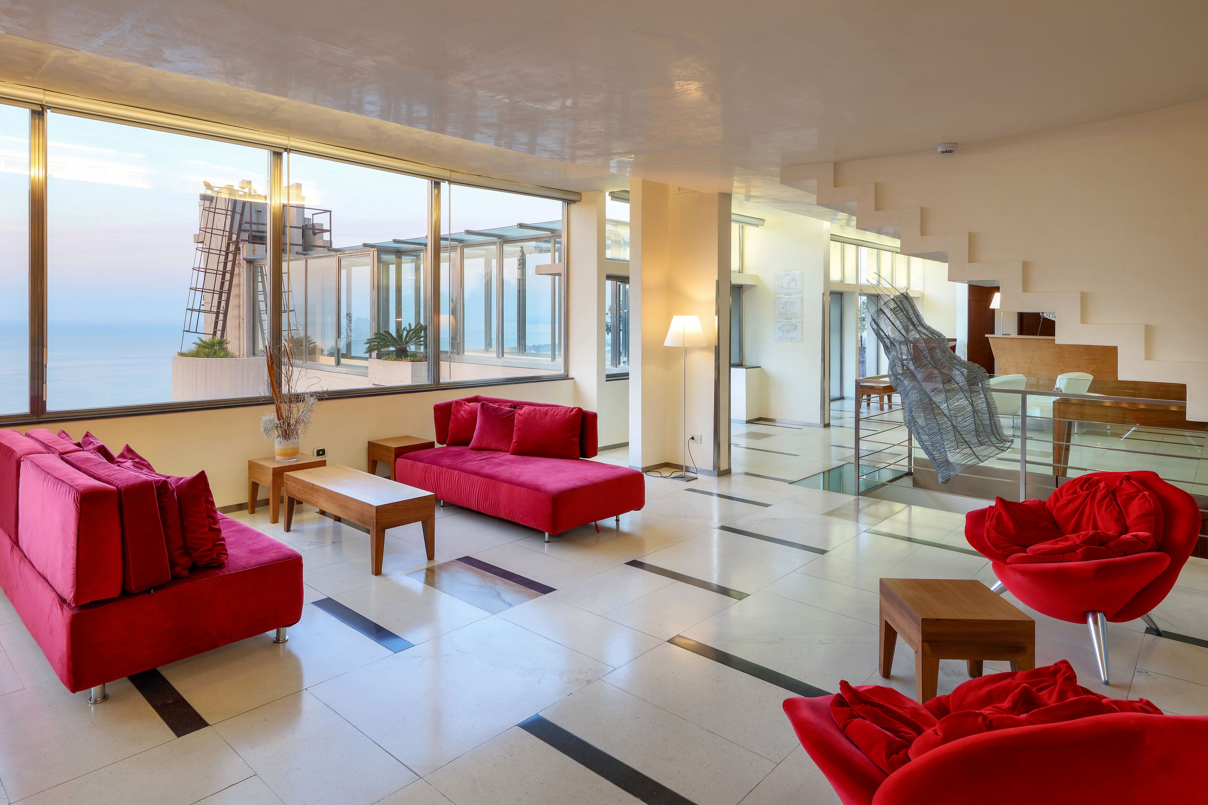 Spacious contemporary living room features bold red sofas and armchairs arranged around light wooden tables. Large floor-to-ceiling windows open to a balcony with ocean views and soft sunset light. The interior includes glossy tiled floors, minimalist staircase, and glass railings that enhance the airy, open feel. Neutral walls and clean lines contrast with the vivid red seating to create a modern, stylish atmosphere.
