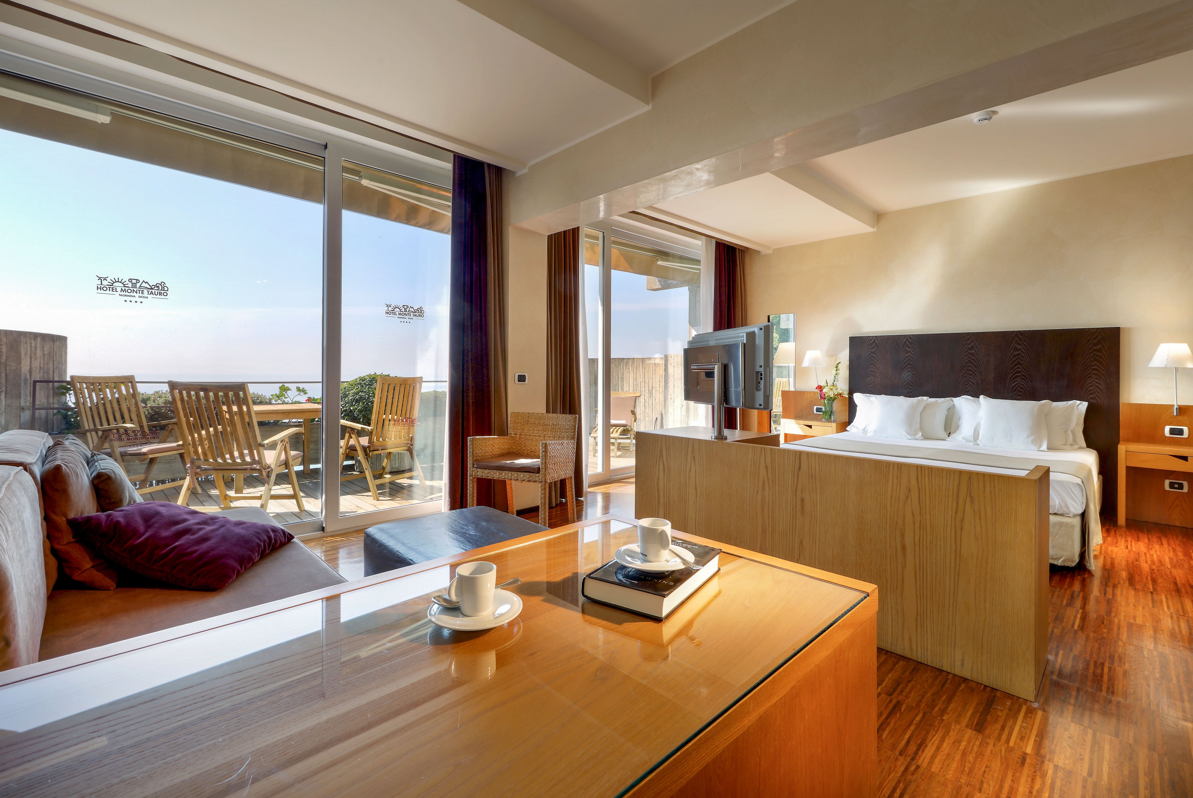 Contemporary hotel suite features a spacious bedroom and living area with warm wooden flooring. Large sliding glass doors open onto a balcony with wooden lounge chairs overlooking the sea and coastline. A sleek desk with coffee cups, saucers, and books sits in the foreground, while a flat-screen TV divides the sleeping and sitting areas. Natural light floods the room, highlighting the minimalist decor and relaxing vacation atmosphere.