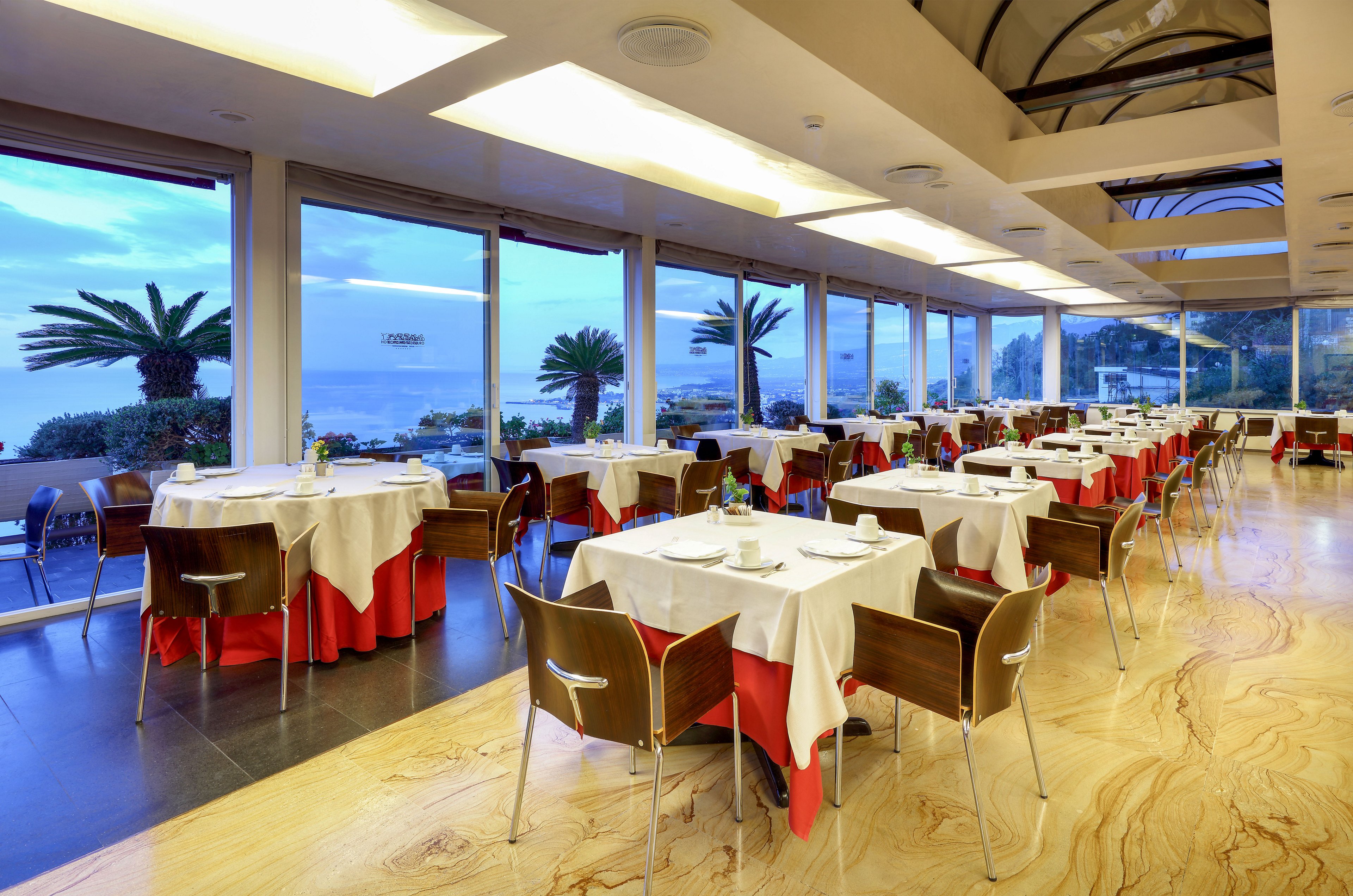 Spacious restaurant dining room with neatly arranged tables overlooks a coastal landscape through large floor-to-ceiling windows. Round tables are covered with white tablecloths and red skirting, each set with simple place settings and glassware. Wooden chairs surround the tables, and the polished floor reflects the warm interior lighting. Outside, palm trees and the sea create a serene backdrop under a soft evening sky.