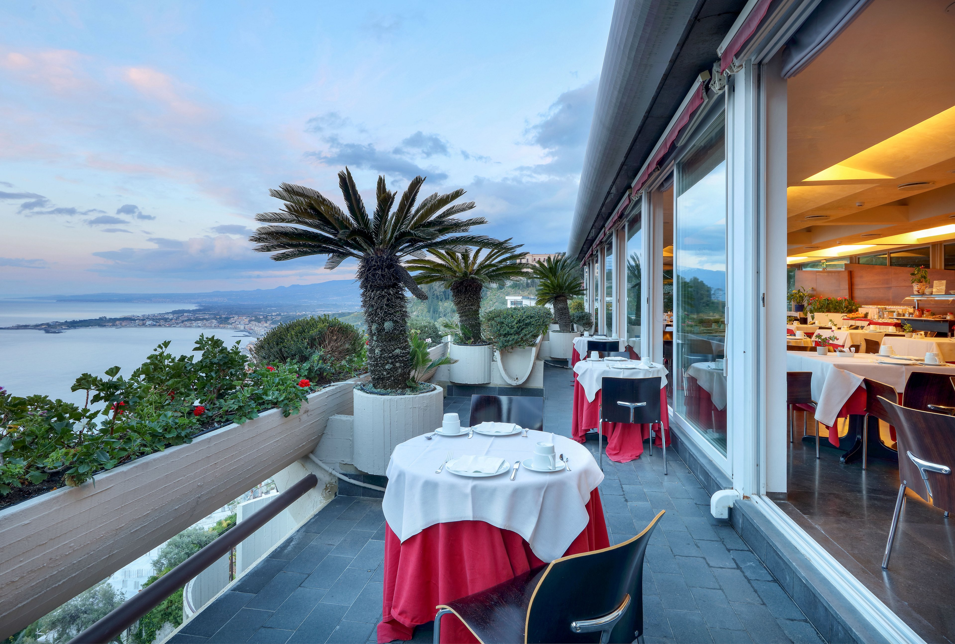 An elegant restaurant terrace overlooks a coastal landscape at dusk. Tables are set with white cloths and red table skirts, ready for guests beside glass windows revealing the indoor dining area. Potted palm trees and lush greenery line the balcony, framing the expansive ocean and shoreline view. Soft evening light and pastel sky tones create a relaxed, romantic atmosphere.