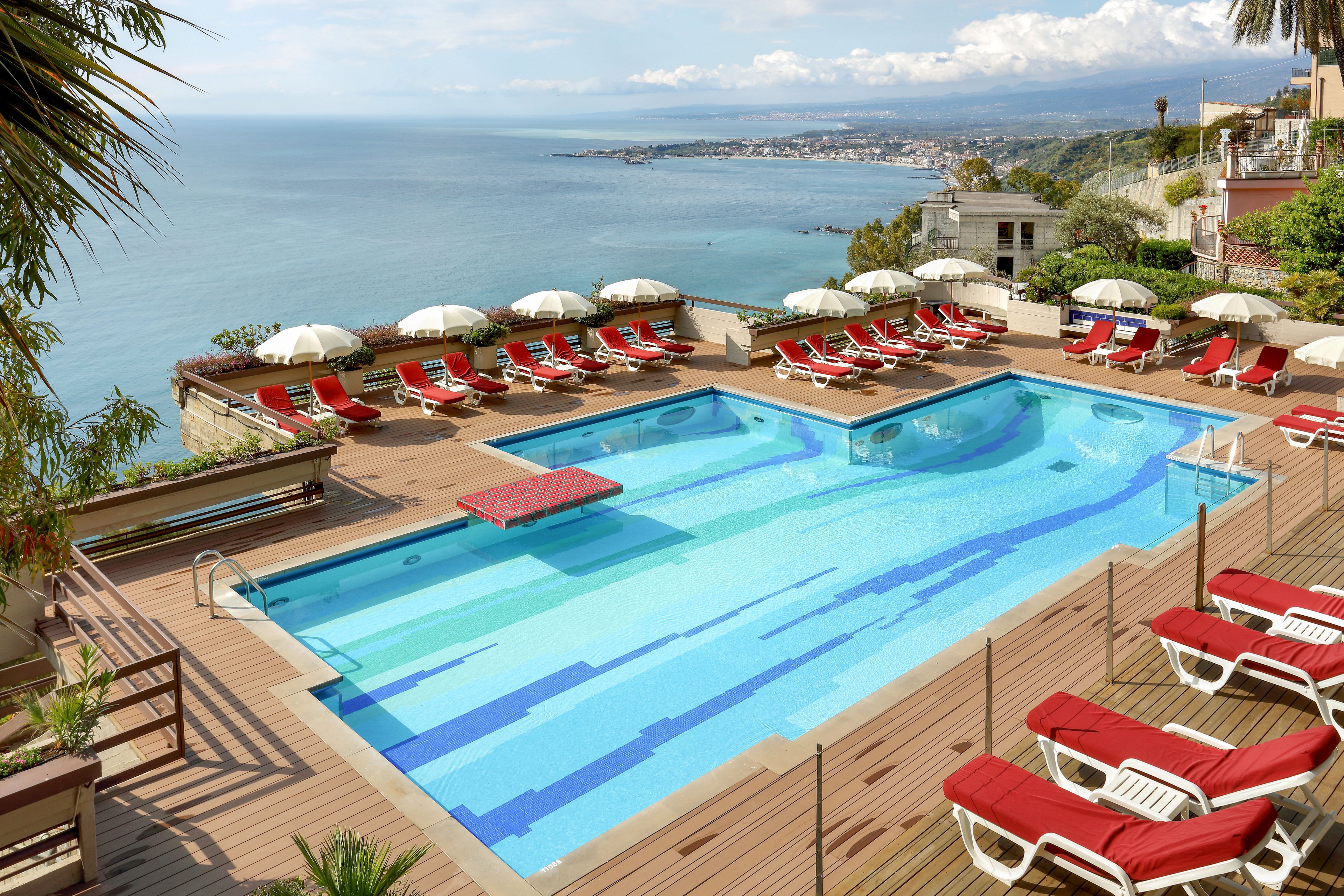 An elevated hotel pool terrace overlooks a sweeping coastal landscape and calm blue sea. The scene features a large central swimming pool with adjacent smaller pools, surrounded by red-cushioned sun loungers and white umbrellas. Wooden decking and potted greenery frame the water, while buildings and hills stretch along the shoreline in the distance. Bright daylight and clear skies create a relaxed, vacation atmosphere.