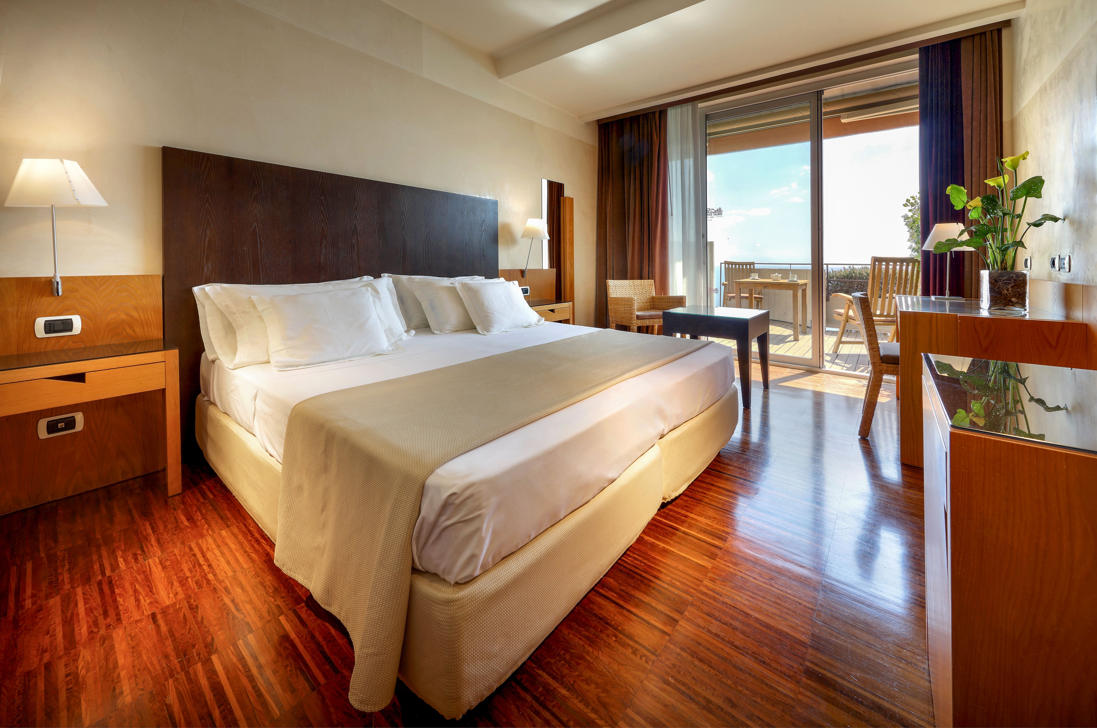 A contemporary hotel room features a large double bed with crisp white linens and wooden headboard. Matching bedside tables, a desk, and a small seating area complement the polished wooden floor. Sunlight streams in through wide glass doors that open onto a balcony with outdoor chairs and a scenic view. Warm lighting and neutral tones create a cozy, inviting atmosphere suitable for relaxation or business travel.