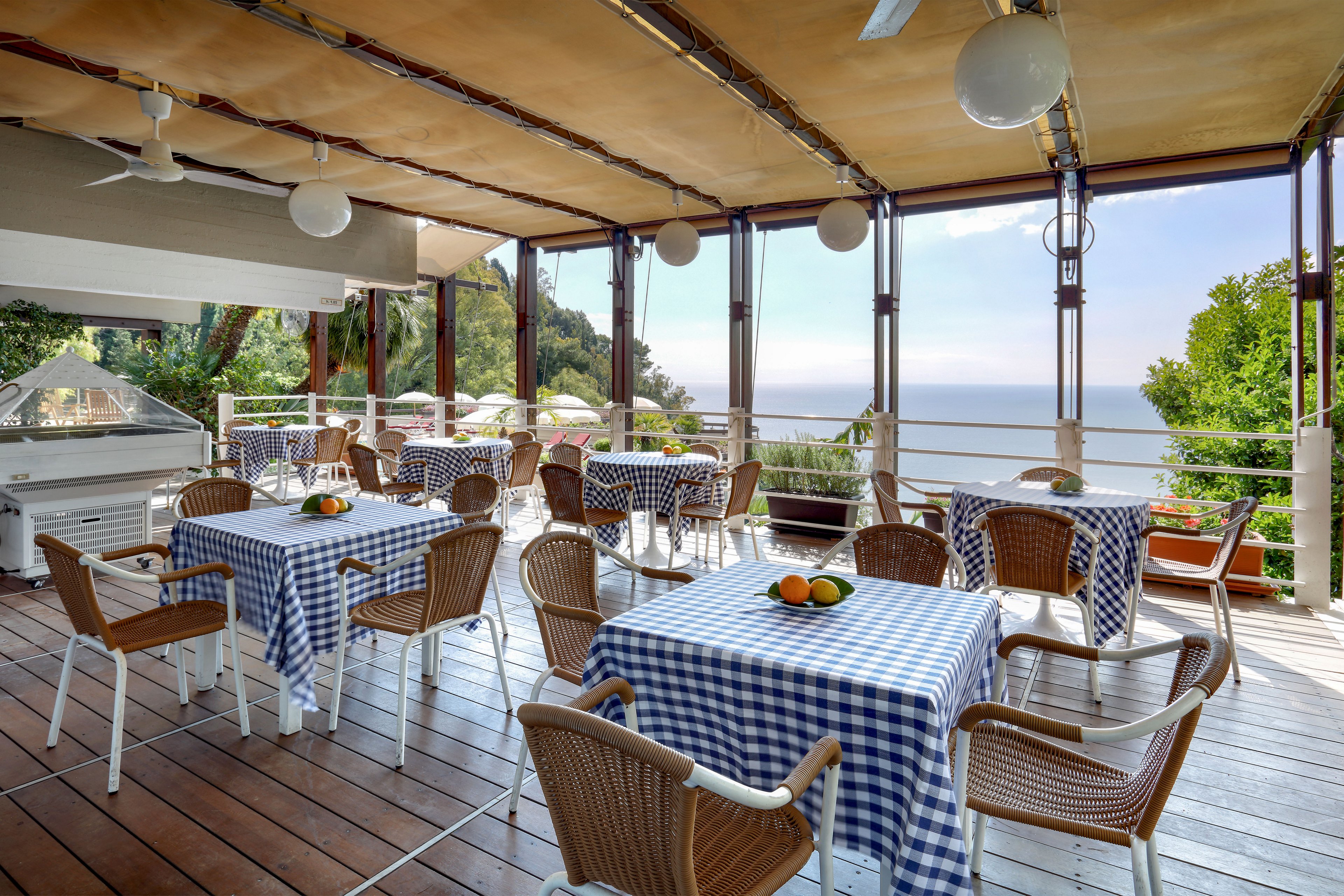 Open-air terrace dining area overlooking a calm sea and distant horizon. Several tables are covered with blue and white checkered tablecloths and surrounded by wicker-style chairs. Bowls of fresh citrus fruit sit on the tables as simple centerpieces. The space is shaded by a canopy roof, with railings framing lush greenery and umbrellas below.