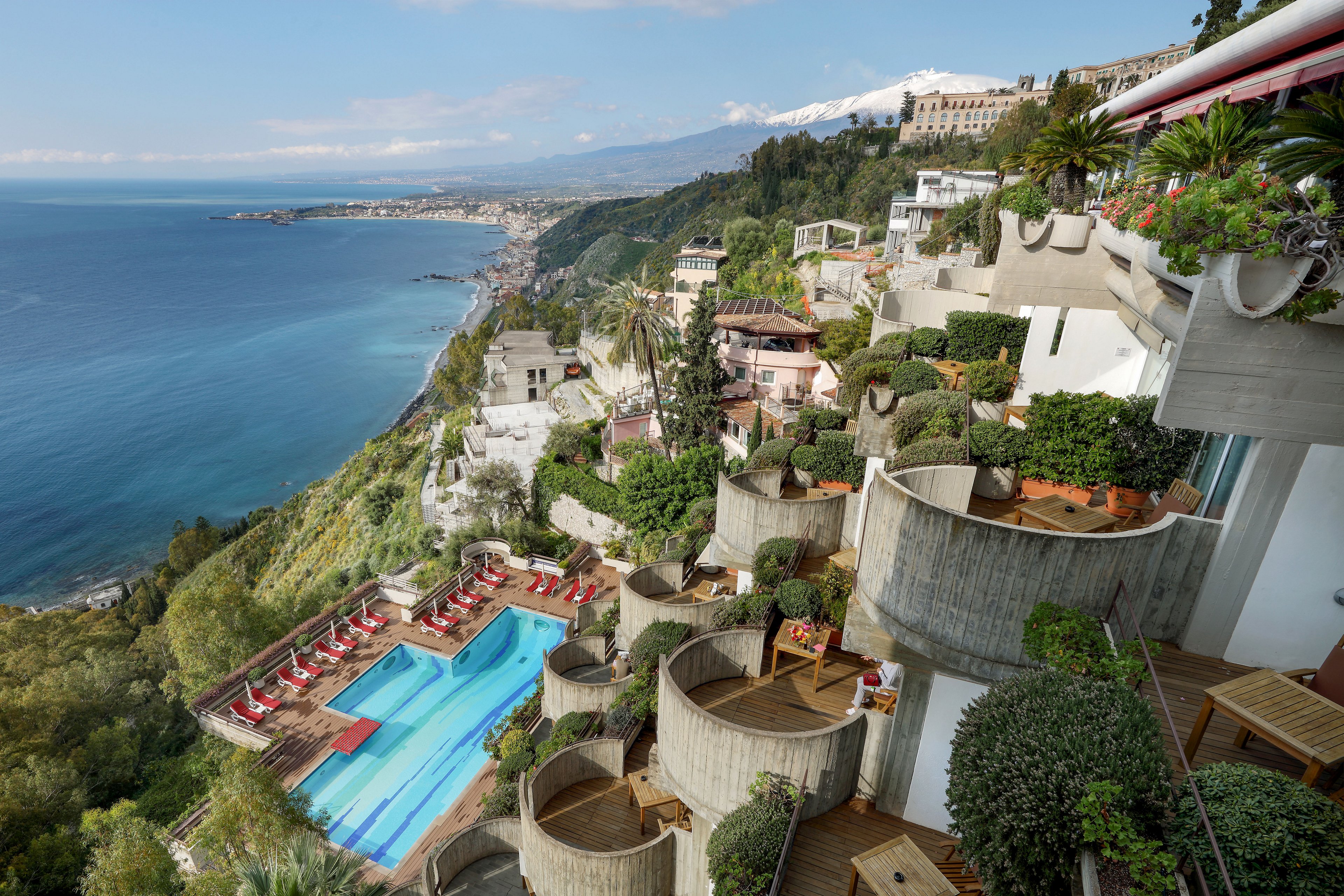 A luxury resort is built on terraced levels overlooking a vast blue coastline. Curved concrete balconies and lush greenery cascade down the hillside, framing an elongated outdoor swimming pool with red sun loungers. The scene captures a dramatic drop to the sea, distant town structures, and mountains under a hazy sky. The overall atmosphere is tranquil and scenic, emphasizing leisure and panoramic ocean views.