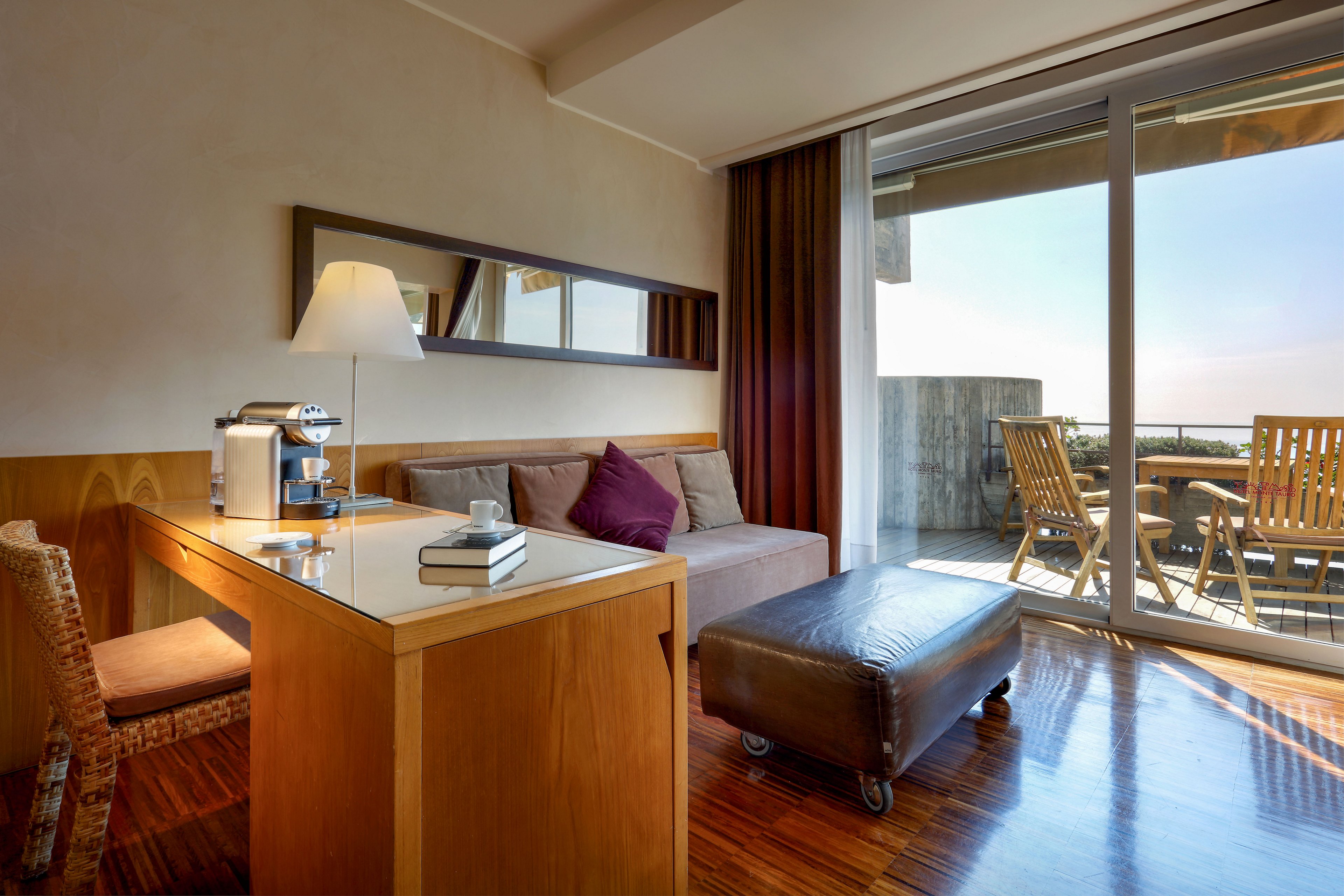 Contemporary hotel room features a cozy seating area with a sofa, ottoman, and wooden desk. A coffee machine and cup sit on the desk beside a book, adding a sense of comfort and relaxation. Large sliding glass doors open to a balcony with wooden chairs and table, overlooking a bright outdoor view. Warm natural light and polished wooden floors create an inviting, tranquil atmosphere.