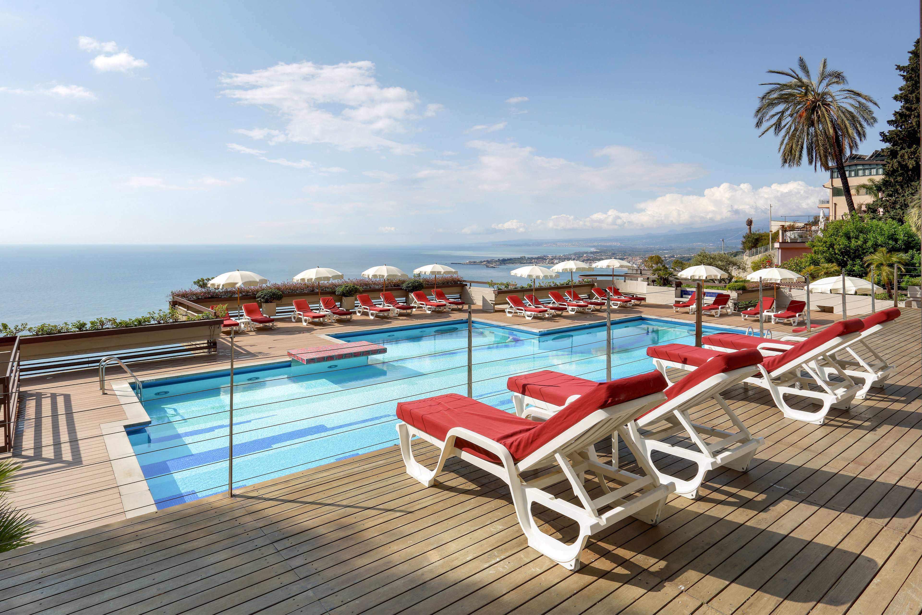 An elevated outdoor swimming pool terrace overlooks a calm sea and distant coastline. Red-cushioned white sun loungers line the wooden deck around the clear blue pool. White umbrellas and additional seating are arranged neatly, suggesting a relaxing resort environment. A tall palm tree and scattered buildings frame the scene under a bright, partly cloudy sky.