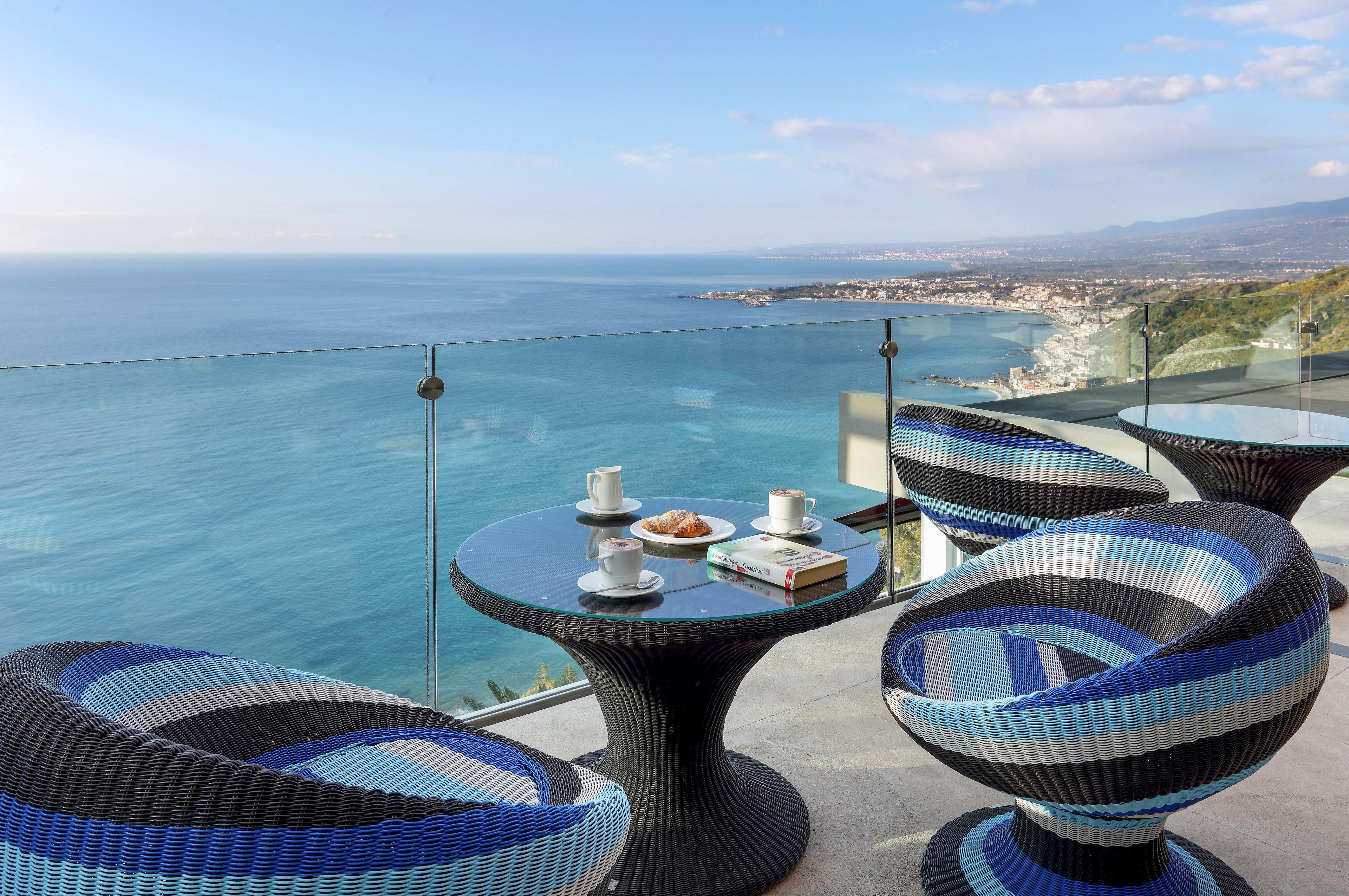 A modern terrace features round wicker chairs and a small glass-top table overlooking a vast blue ocean. On the table are cups of coffee, a plate of pastries, and a book, suggesting a relaxed café setting. Glass railings frame the edge of the balcony, opening to a panoramic view of the coastline and distant town. Soft daylight and clear skies create a serene, leisurely atmosphere.