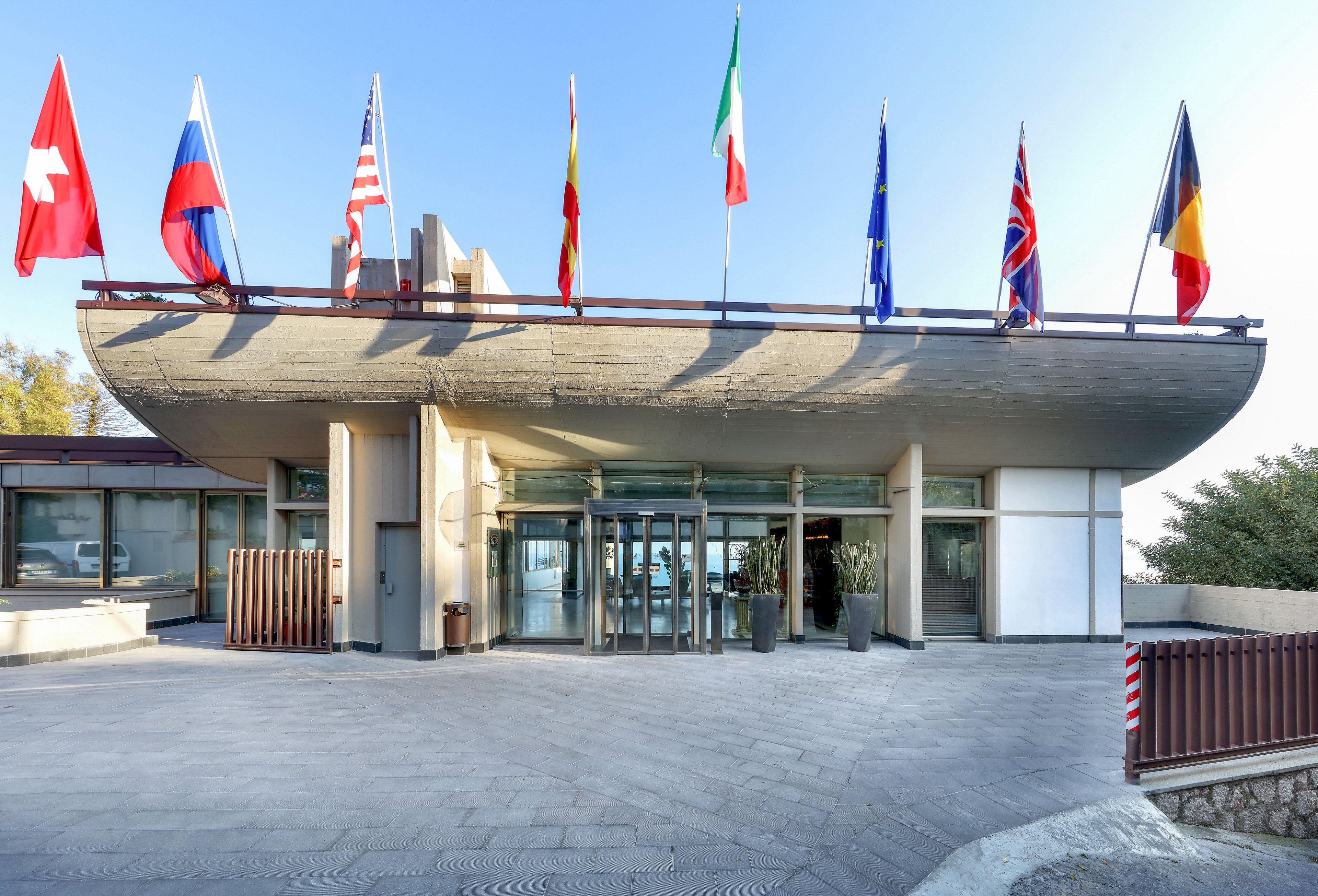 A contemporary building entrance features a wide glass doorway beneath a curved concrete overhang. Multiple national flags are mounted along the roofline, adding color against a clear blue sky. The foreground shows a clean, paved plaza leading up to the structure, with metal railings and planters accenting the space. The scene is brightly lit, suggesting a sunny day and a formal, institutional setting.