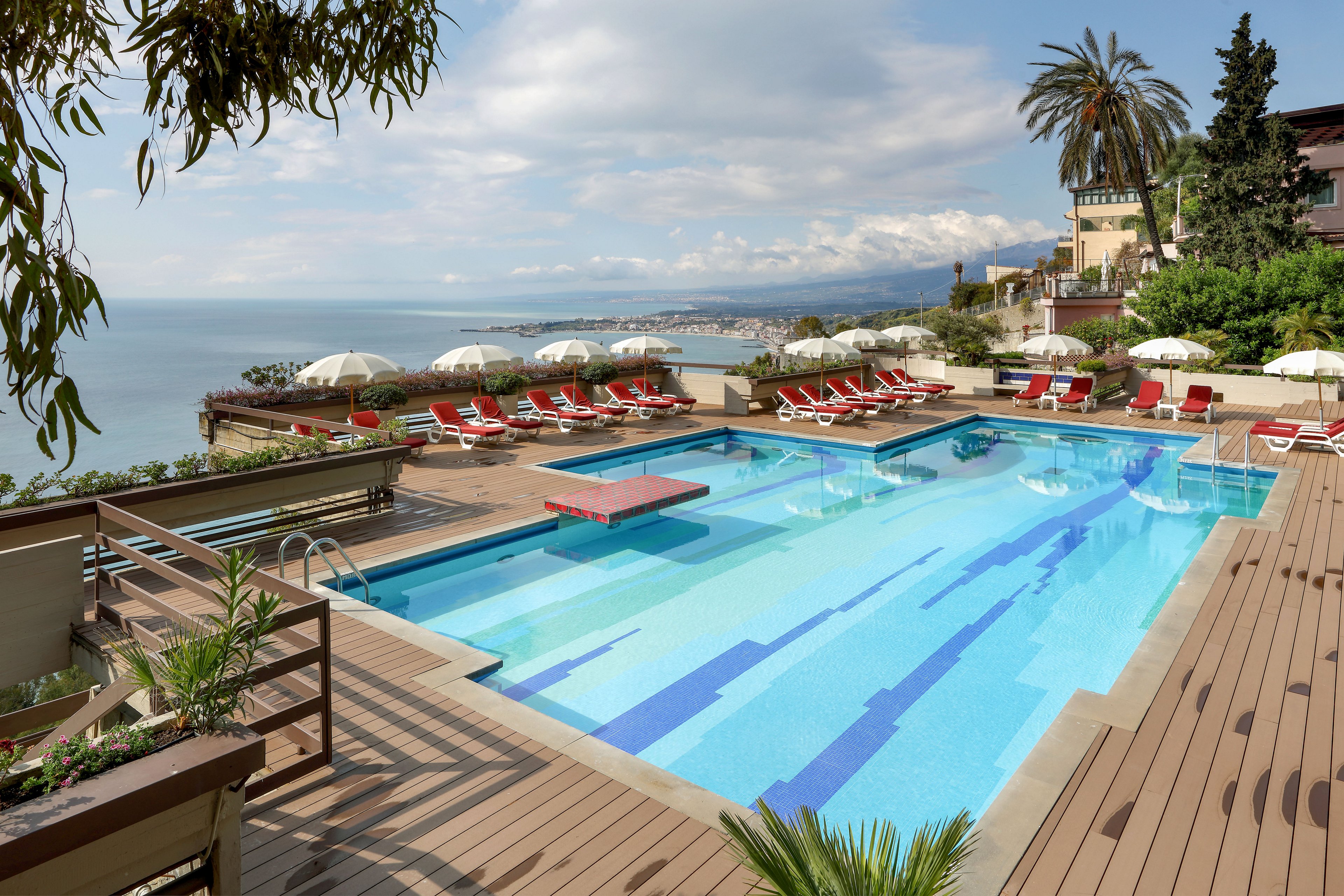 An elevated outdoor swimming pool overlooks a calm sea and distant coastline. Red sun loungers and white umbrellas line the wooden deck around the pool, creating a resort-style setting. Palm trees, lush greenery, and nearby buildings frame the terrace, while soft clouds hang over the horizon. The scene suggests a relaxing vacation atmosphere with clear water and ample seating for guests.