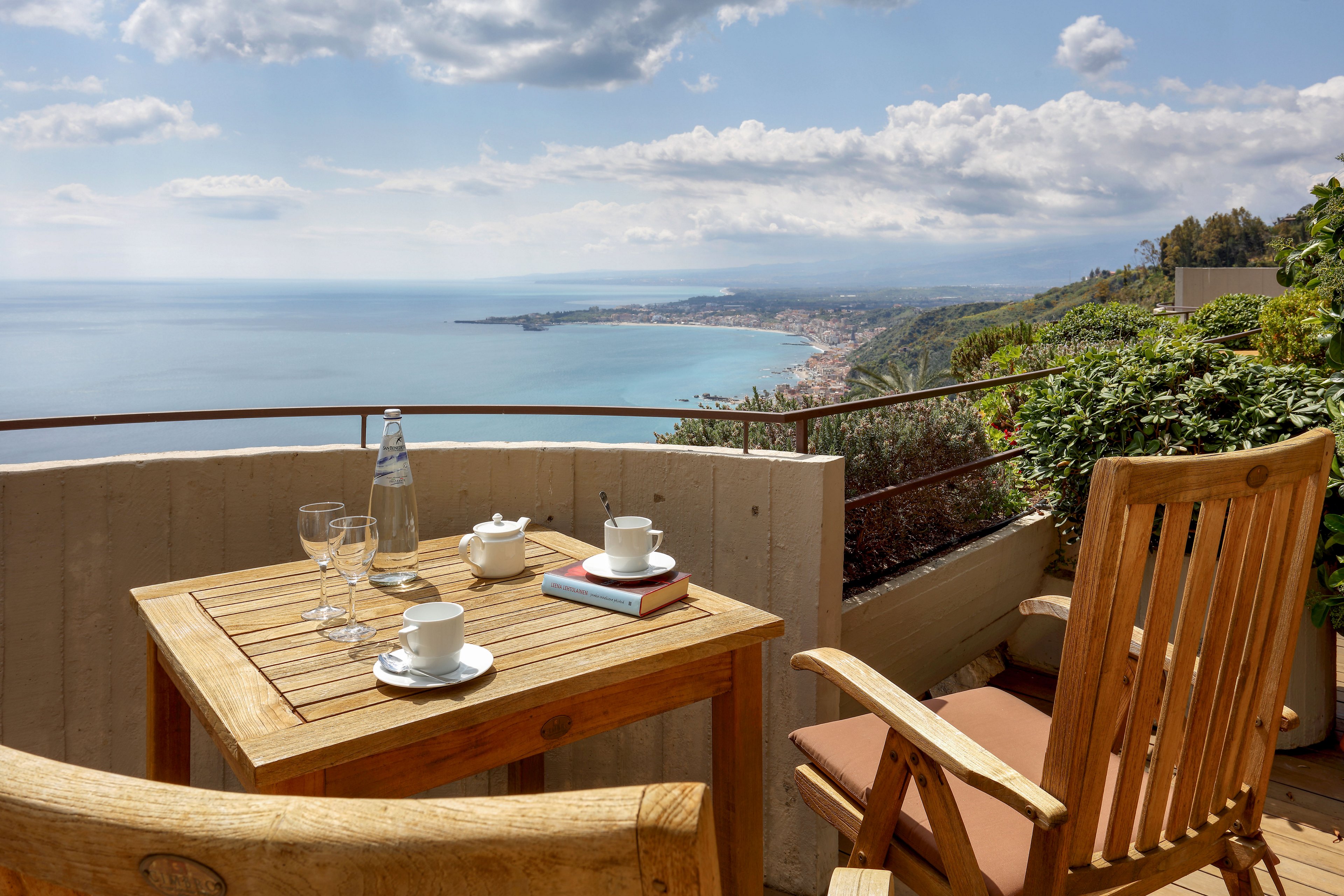 A wooden terrace table is set with coffee cups, glasses, and a glass bottle overlooking a vast coastal landscape. Wooden chairs surround the table, inviting relaxation on the balcony. The scene captures a sweeping view of the sea, shoreline, and distant town under a partly cloudy sky. Lush greenery frames the balcony, enhancing the tranquil and leisurely atmosphere.