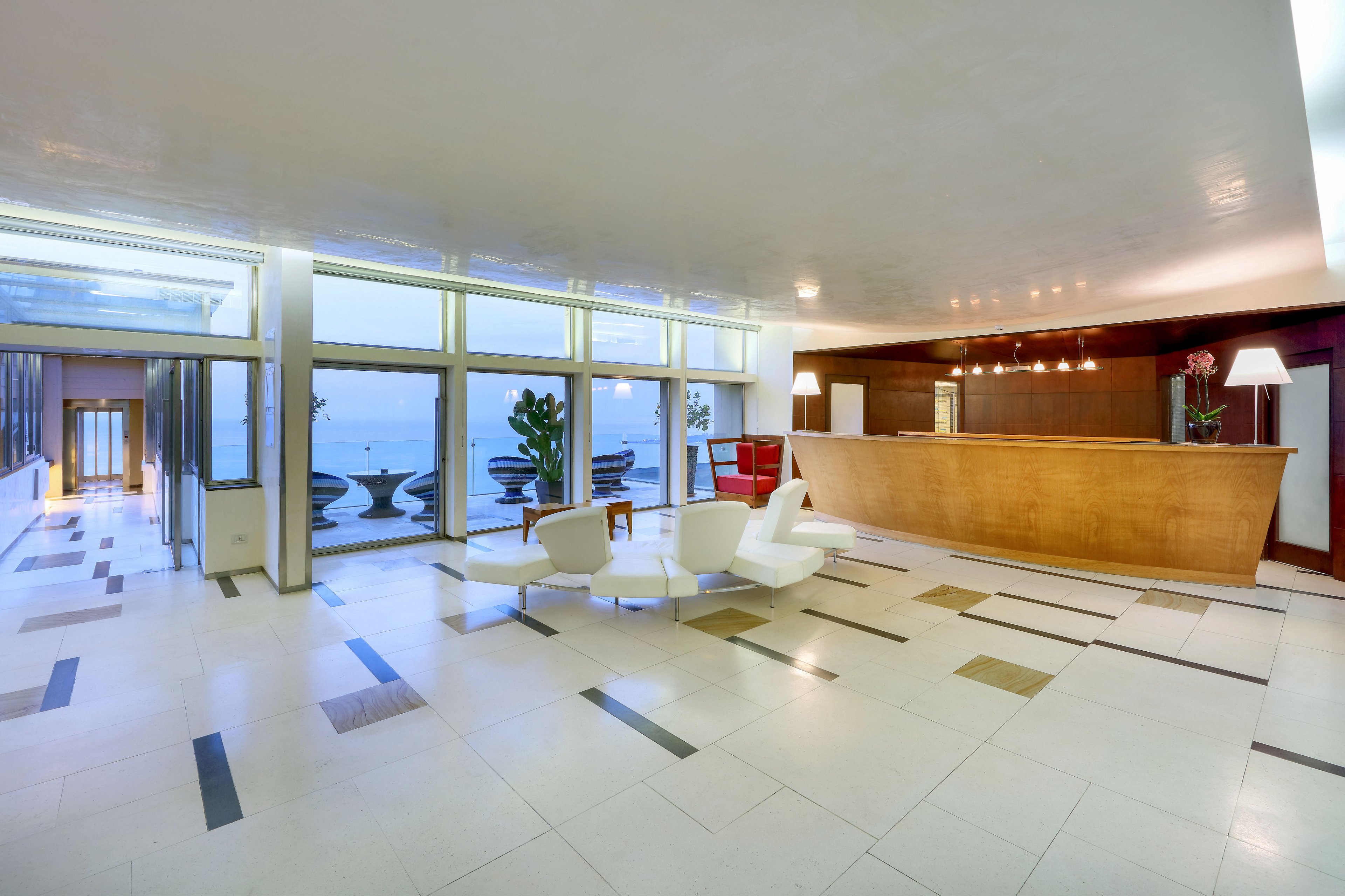 A spacious, contemporary hotel lobby features a long wooden reception desk and sleek white seating. Large floor-to-ceiling windows reveal an outdoor terrace and a distant ocean view. The interior is bright with polished light-colored flooring accented by dark inlays and minimalist decor. Warm lighting above the desk contrasts with the cool natural light from the windows, creating a welcoming atmosphere.