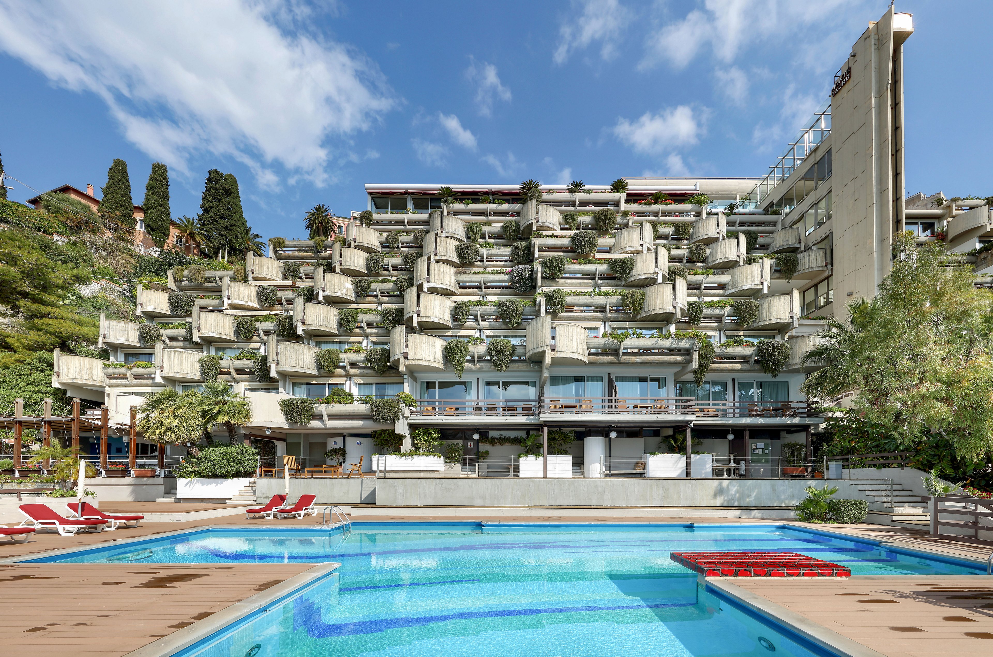 A large terraced hotel building overlooks an outdoor swimming pool in a coastal setting. The architecture features stacked balconies with abundant greenery and plants. Lounge chairs and umbrellas are arranged around the pool deck, suggesting a relaxing vacation atmosphere. Trees and hillside vegetation frame the scene under a bright blue sky with scattered clouds.