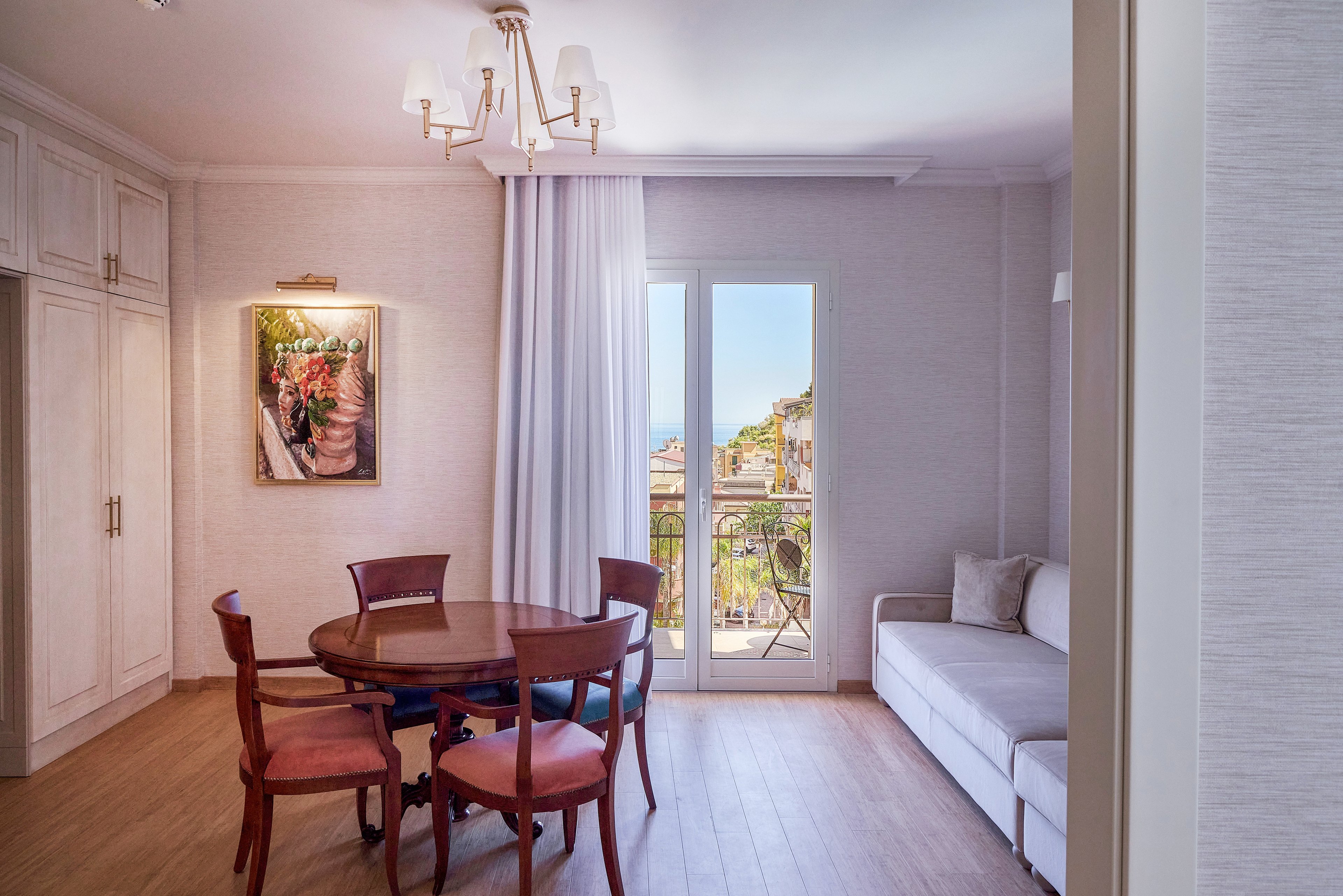 A cozy apartment interior features a round wooden dining table with four chairs beside a wall-mounted painting. Large glass doors open to a balcony overlooking a scenic town and hillside. A light-colored sofa sits along the right wall, and built-in cabinets line the left side of the room. Soft lighting and neutral tones create a warm, inviting atmosphere.