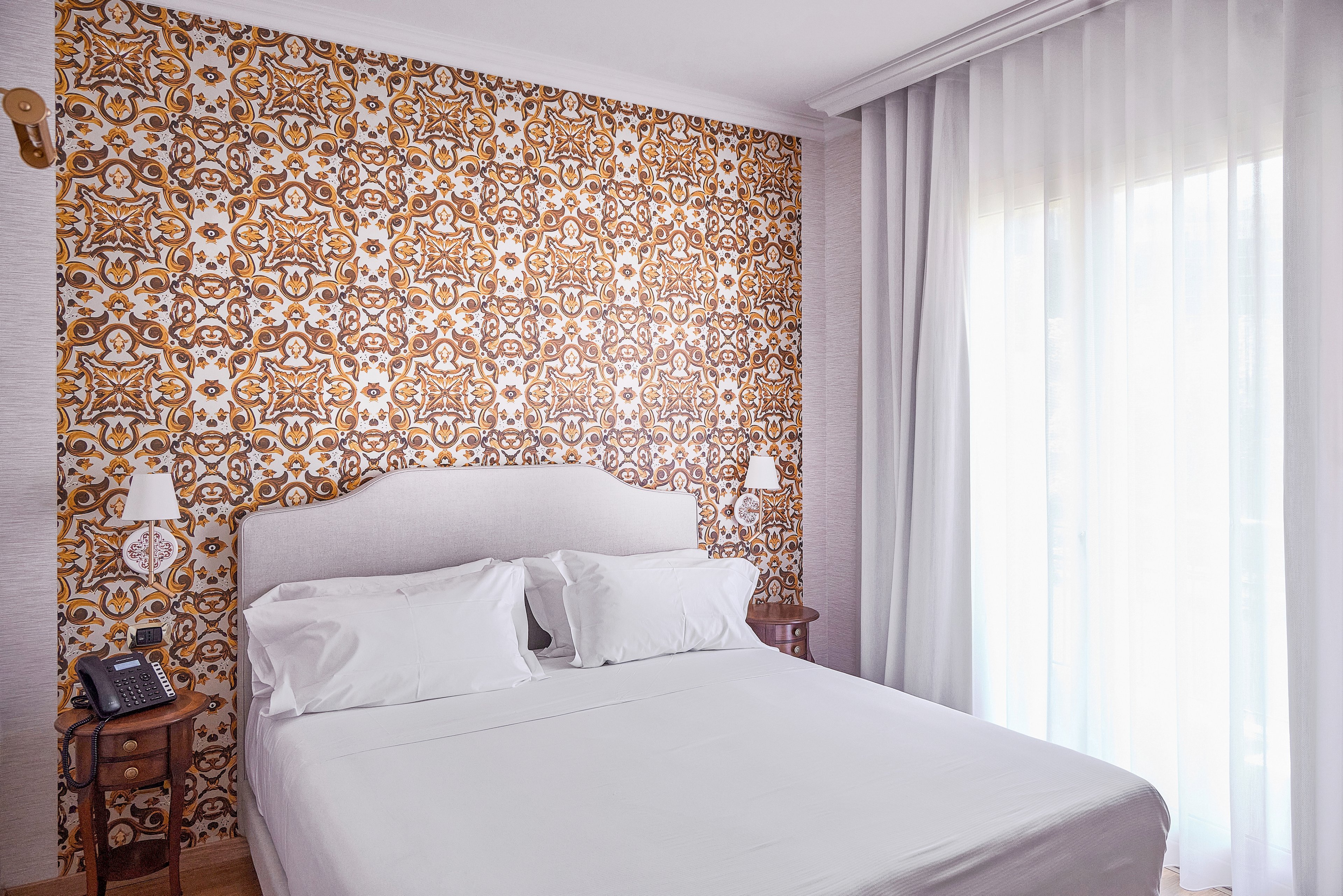 A neatly made double bed with white linens stands against a decorative patterned accent wall in a bright bedroom. Two small wooden bedside tables flank the bed, each with a wall-mounted reading lamp, and one holding a telephone. Sheer white curtains cover a large window, allowing soft natural light to fill the room. The overall scene feels clean, inviting, and contemporary with a touch of vintage style.