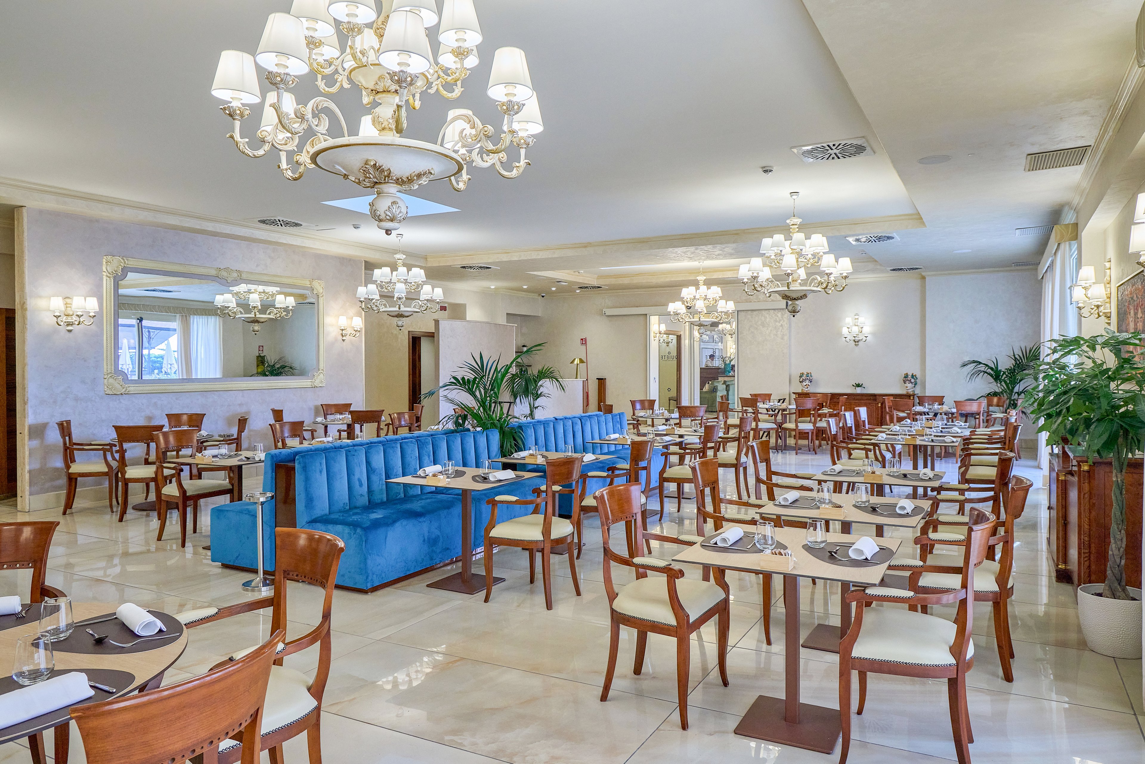 Spacious restaurant interior featuring neatly arranged wooden tables and chairs around a central blue upholstered sofa. Multiple ornate chandeliers hang from the ceiling, casting warm light across the polished floor. Table settings include placemats, napkins, and glassware, suggesting readiness for guests. Large mirrors, potted plants, and soft neutral wall tones contribute to a refined, upscale dining atmosphere.