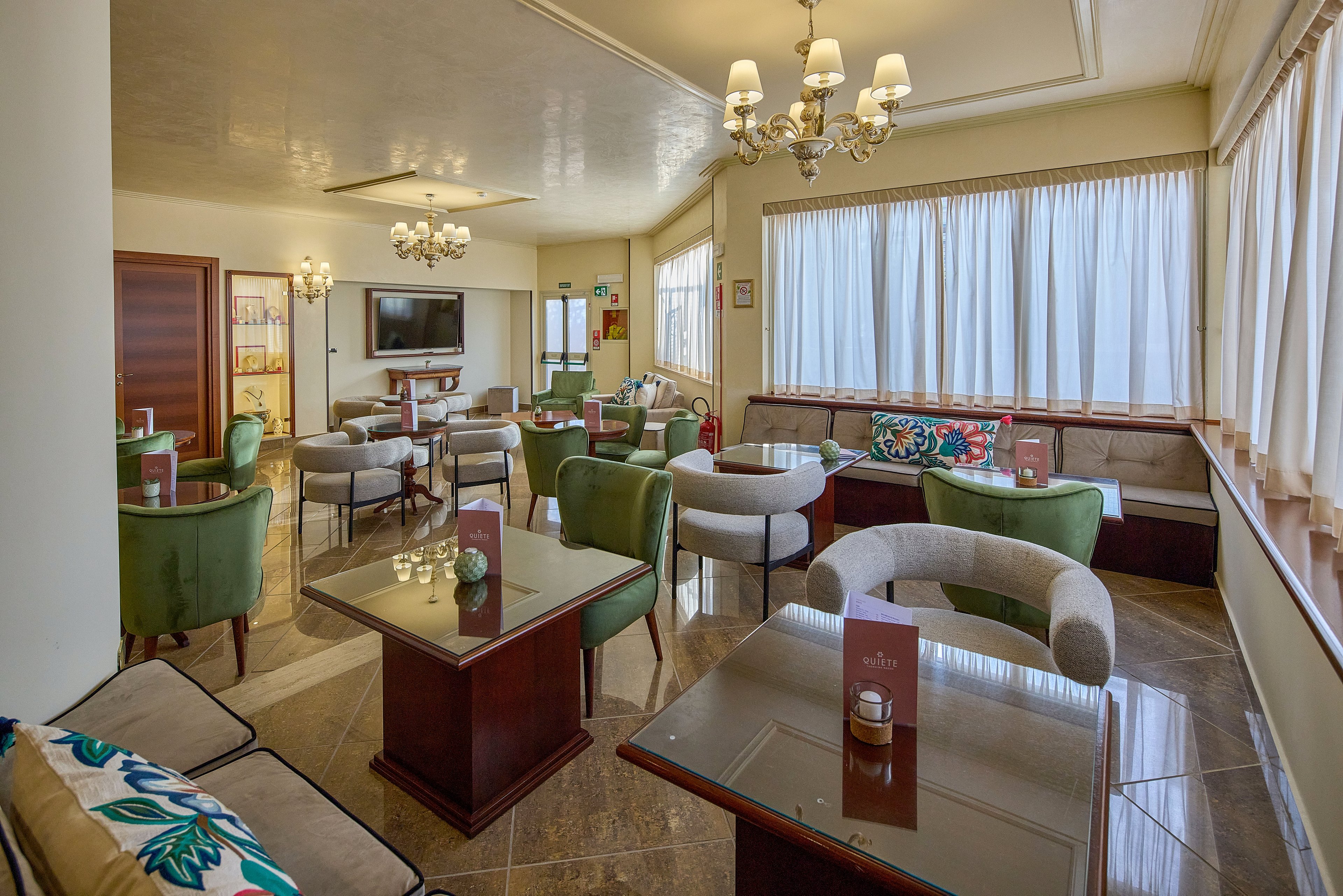 Spacious lounge area with multiple armchairs and sofas arranged around wooden tables. Large windows with sheer curtains fill the room with natural daylight, highlighting the polished floor and neutral walls. Decorative chandeliers hang from the ceiling, and colorful cushions add a touch of vibrancy to the seating. Tabletop menus and candles suggest a relaxed café or hotel bar setting with no people present.