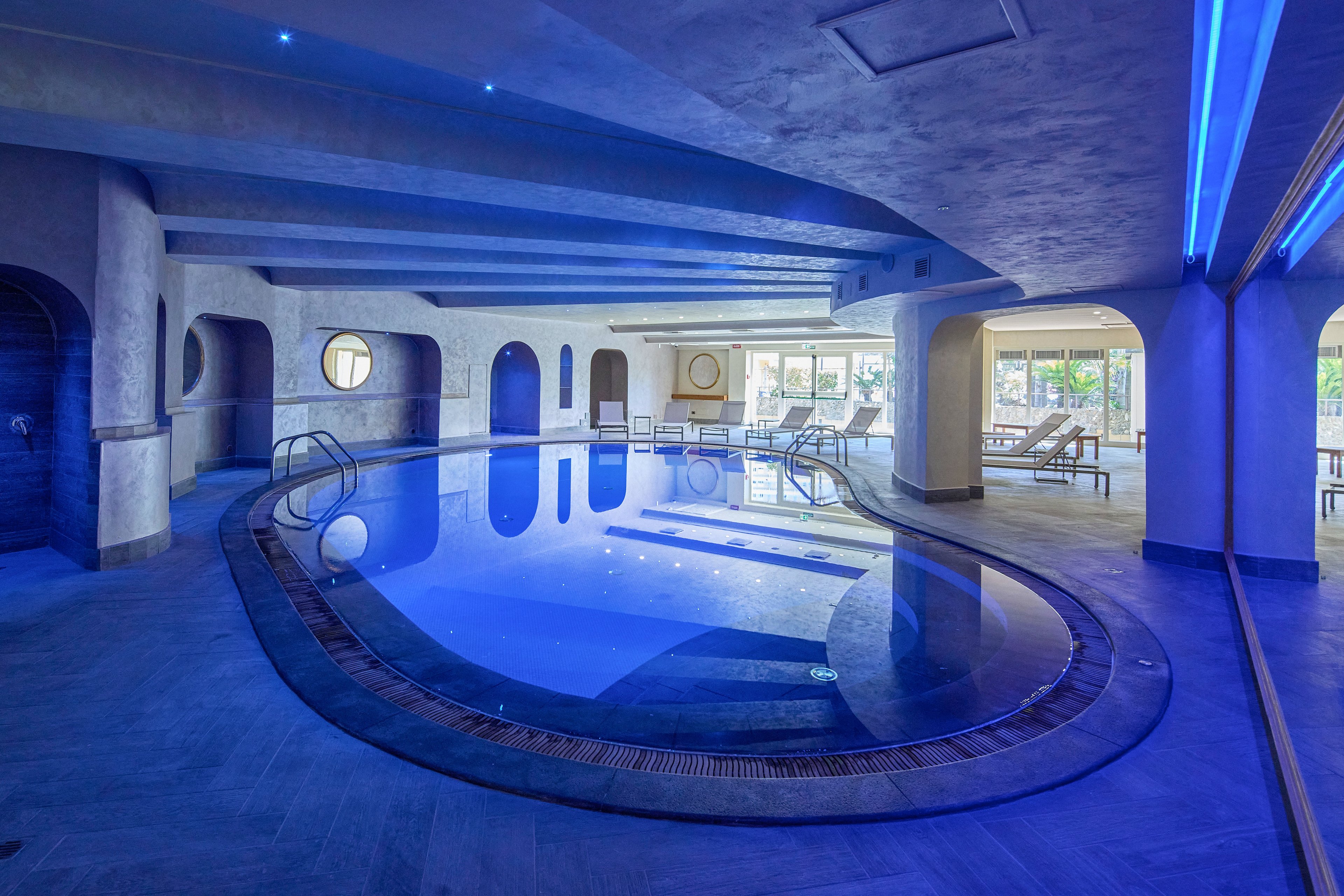Curved indoor swimming pool in a high-end spa or hotel setting. Blue ambient lighting reflects off the water and ceiling, creating a calm, modern atmosphere. Sunlit lounge chairs and large windows are visible in the background, suggesting a relaxing leisure area. Architectural arches and smooth stone flooring add to the sophisticated design.