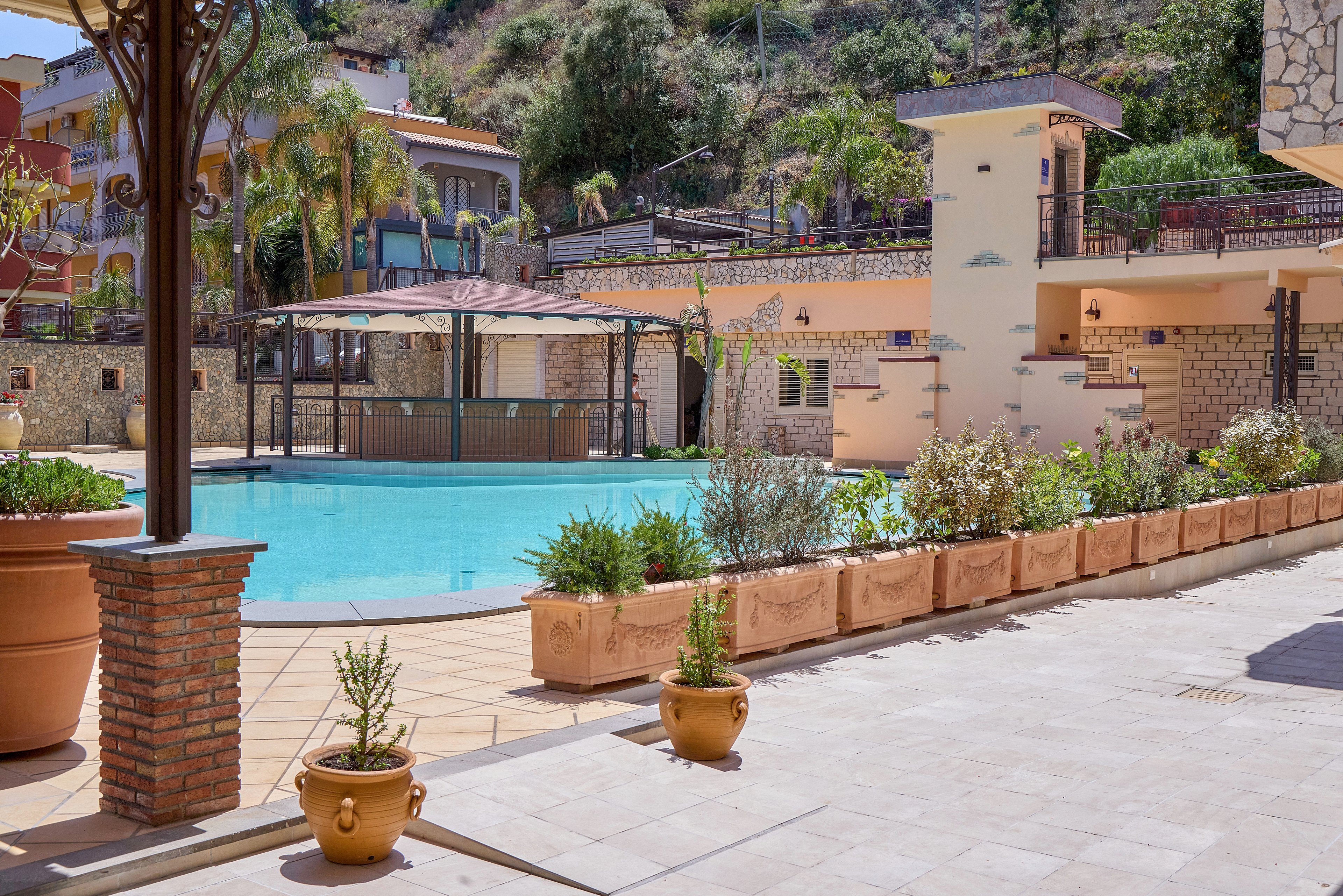 Outdoor swimming pool in a hotel courtyard under bright daylight. A shaded gazebo structure stands over the water, surrounded by tiled deck and potted plants. Terraced buildings and palm trees rise in the background, creating a Mediterranean resort atmosphere. The scene is calm and inviting, with warm stone textures and clear blue water.
