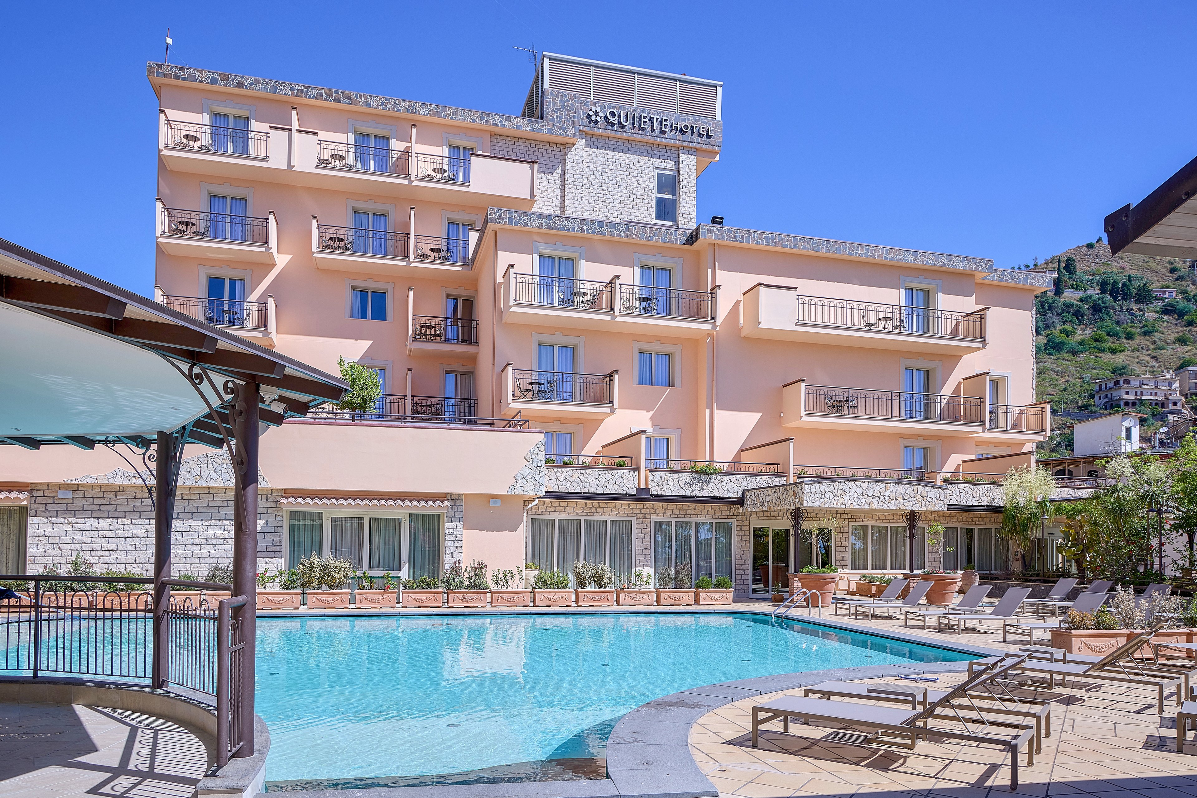 A multi-story peach-colored hotel overlooks a large outdoor swimming pool. Sun loungers and umbrellas line the poolside terrace, creating a relaxing vacation atmosphere. The building features numerous balconies and large windows, with hills and greenery visible in the background. Clear blue sky and bright daylight emphasize the resort’s warm, inviting setting.
